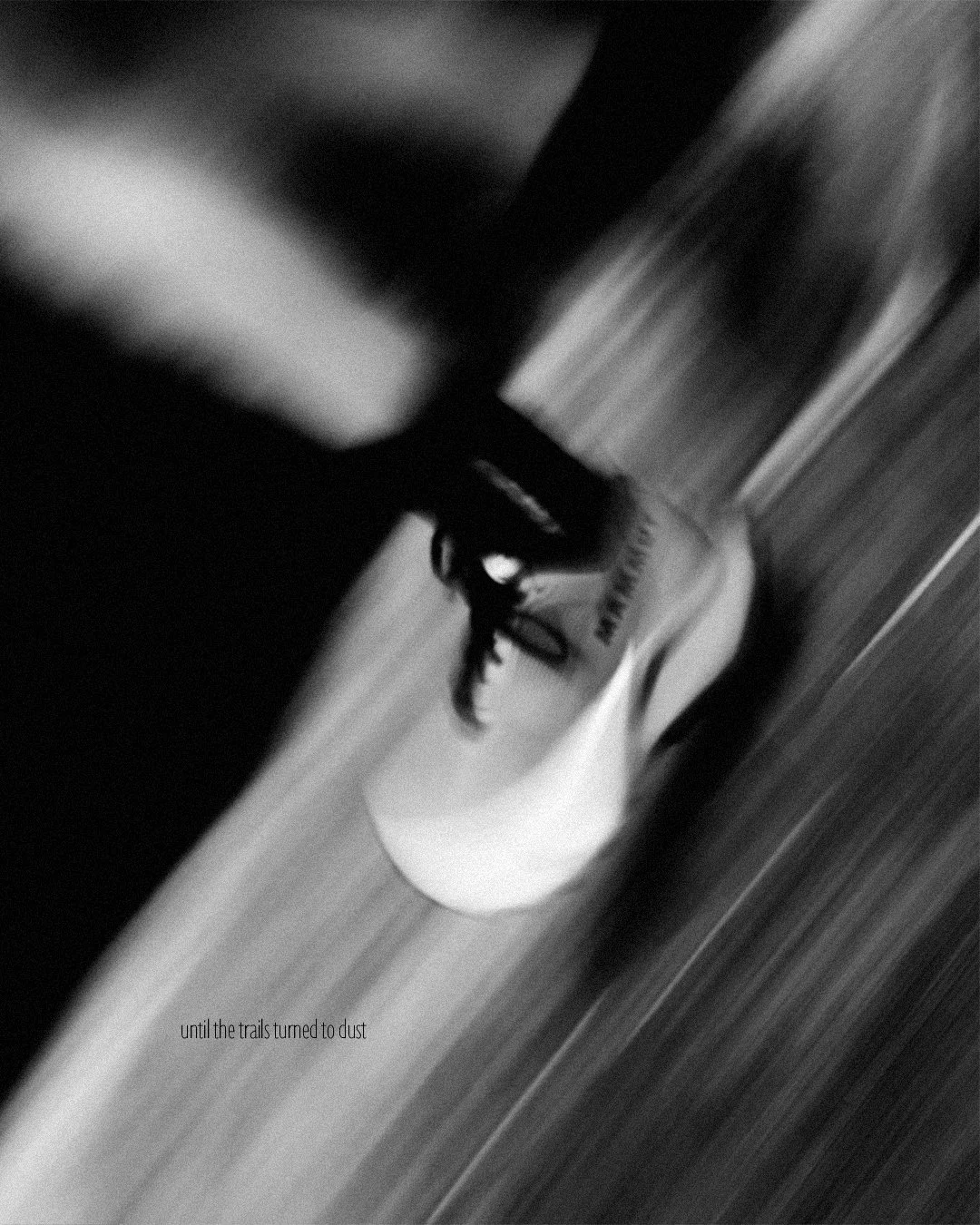 Chaussures de course blanches posées au sol dans une pièce, avec un meuble en arrière-plan. Texte en bas à gauche : 'until the trails turned to dust'