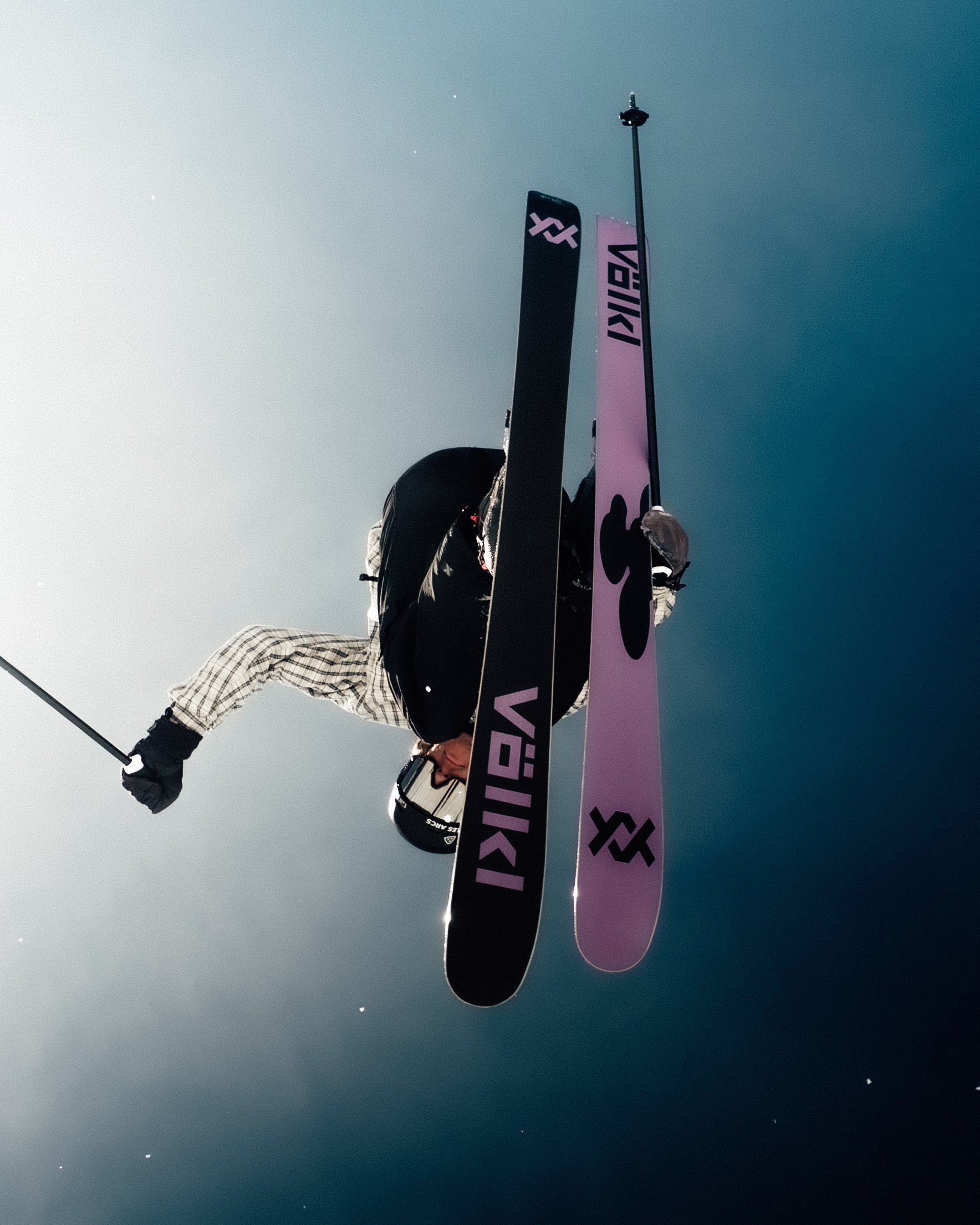Une personne en tenue de ski portant un casque et lunettes, tenant un piolet, debout dans un ciel clair, avec deux skis roses portés verticalement devant elle.