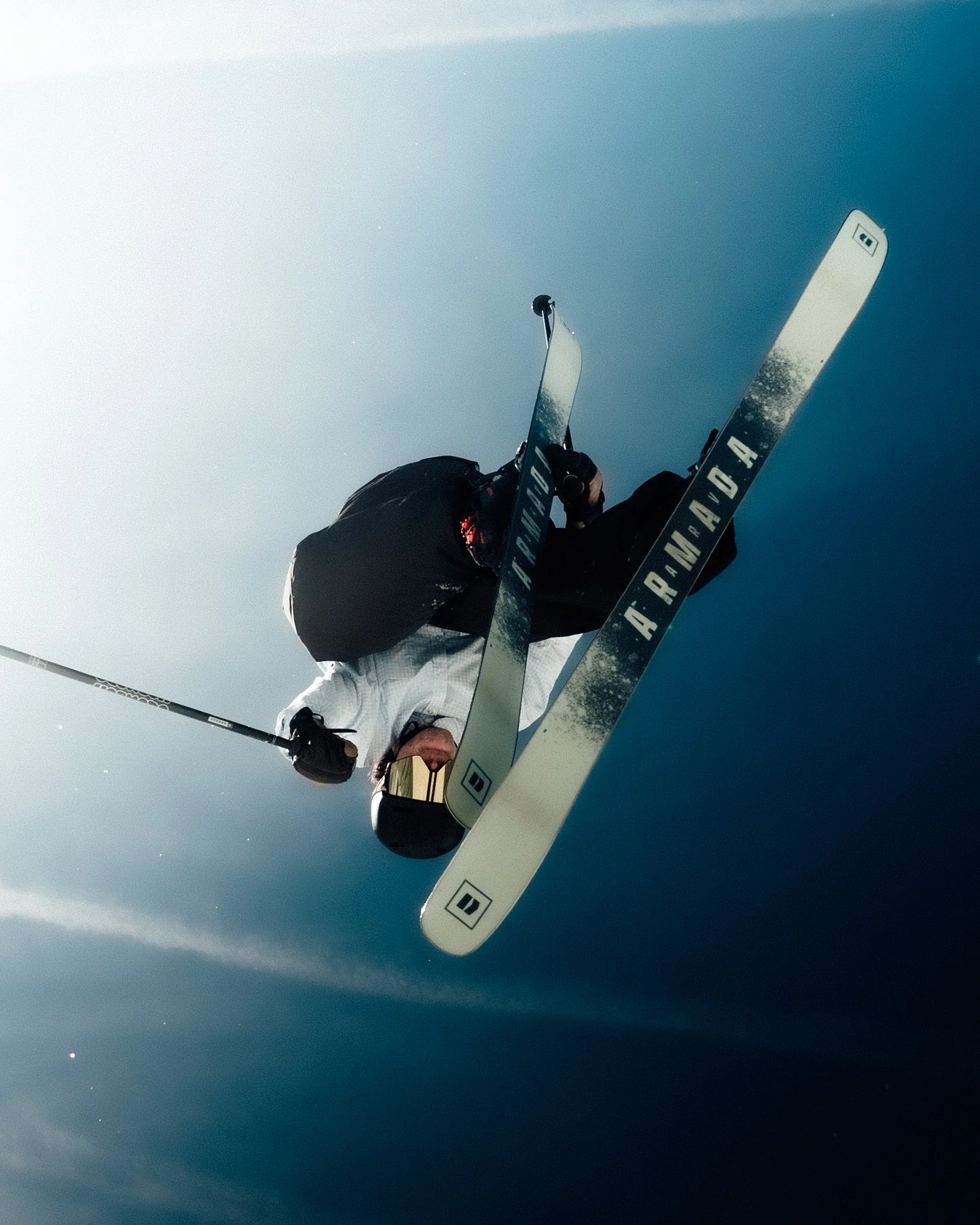 Un skieur portant un casque et des lunettes de ski, en position de descente sur une pente enneigée, tenant deux skis blancs avec des marques en noir. Le ciel est clair et bleu.