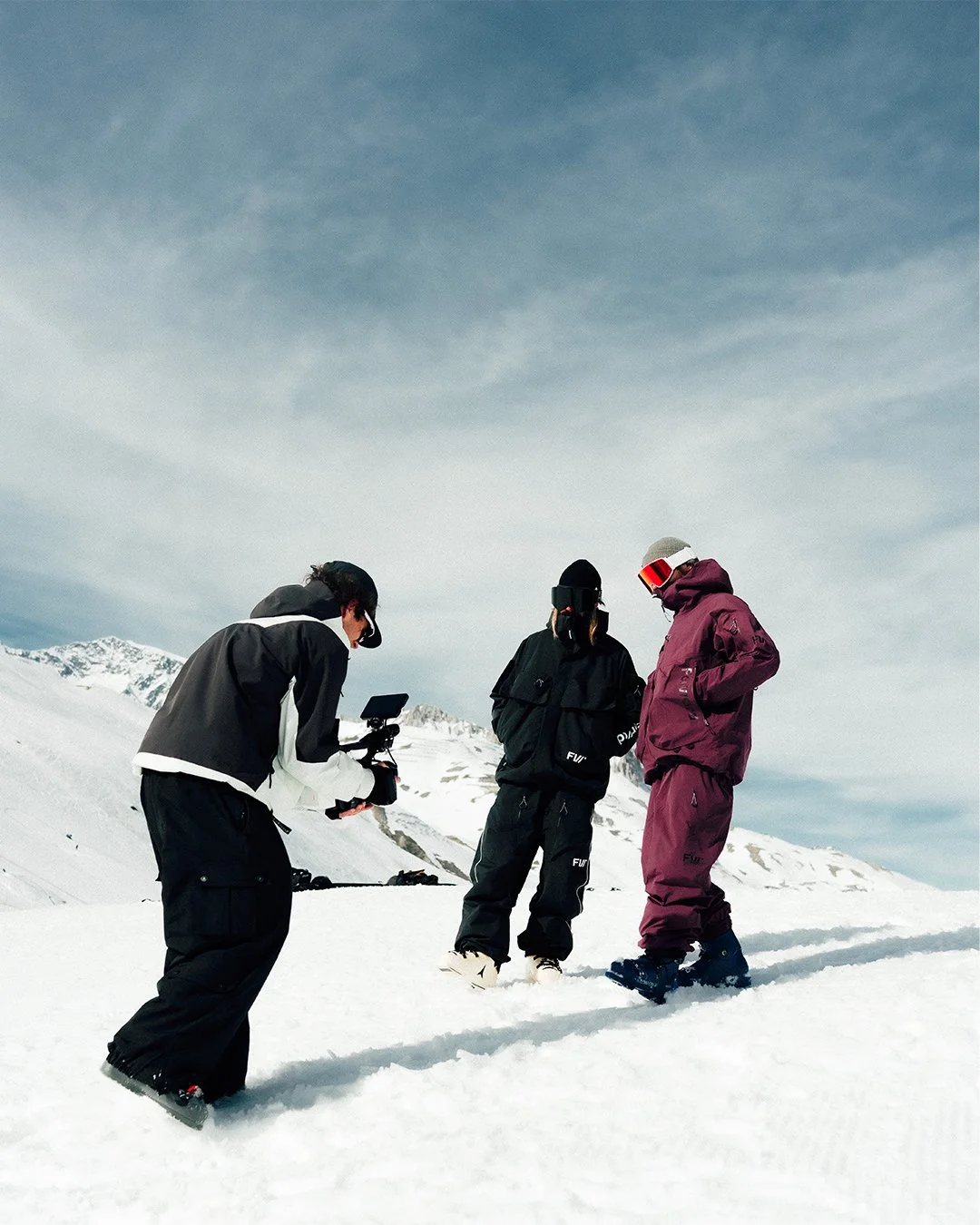 Trois personnes habillées en vêtements de ski sur la neige, une filme une autre personne, probablement pour une vidéo ou une interview, avec un paysage de montagnes enneigées en arrière-plan.