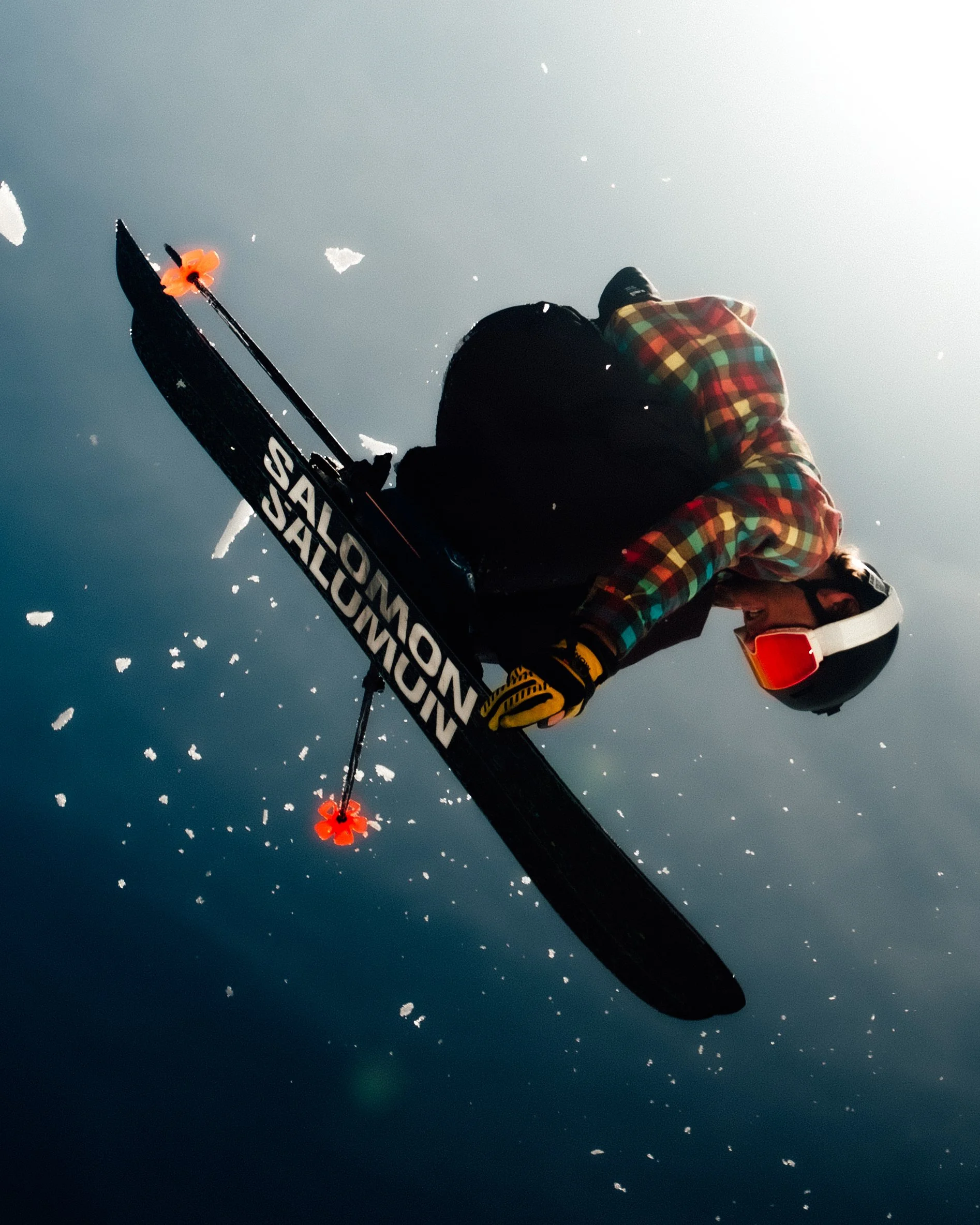 Une personne vêtue d'une veste à carreaux colorés, port du casque de ski, vue de dessous en train de skier sur des montagnes enneigées avec le ciel clair en arrière-plan.