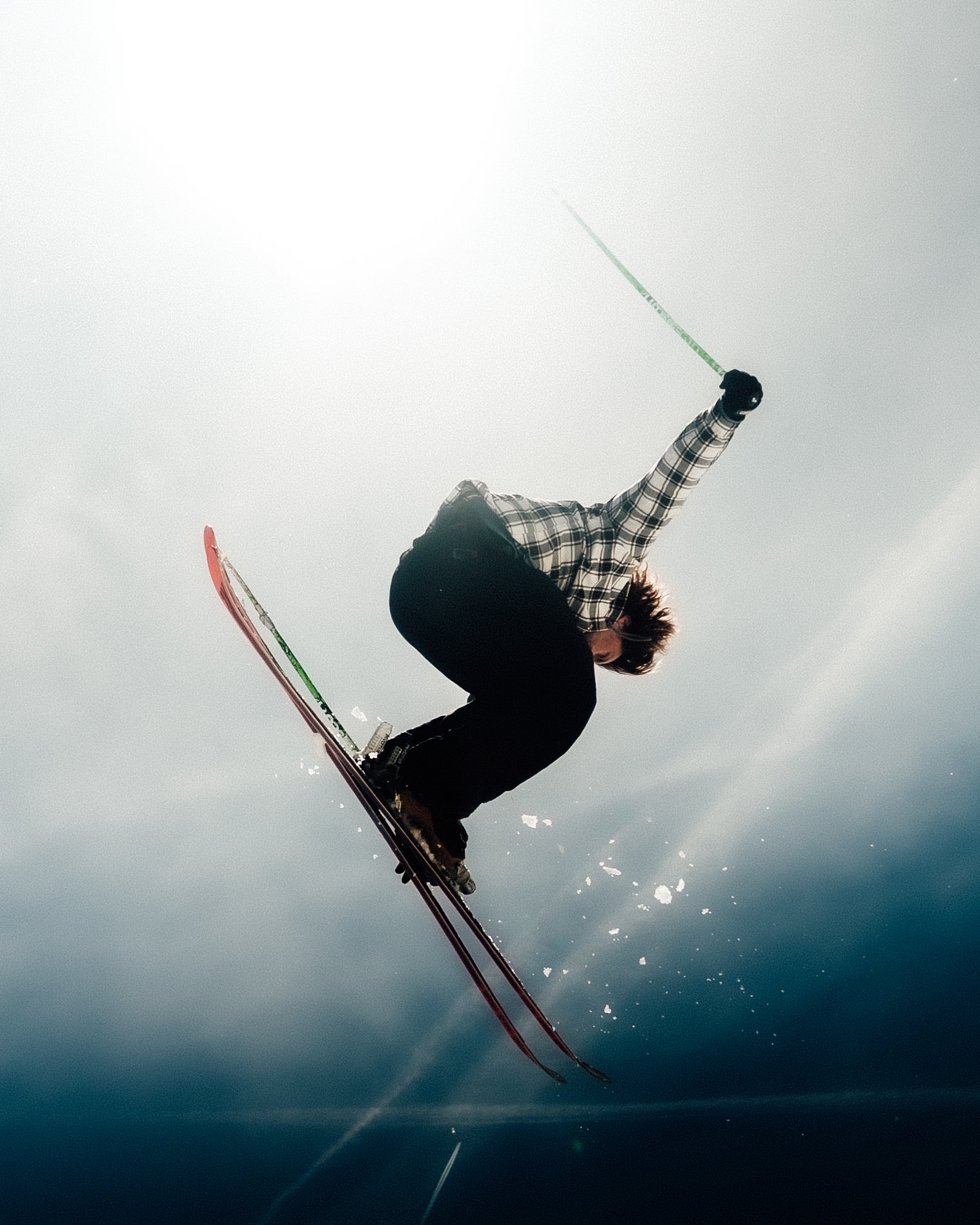 Un skieur en plein saut dans le ciel, vue de dessous, portant un manteau à carreaux et des skis rouges.