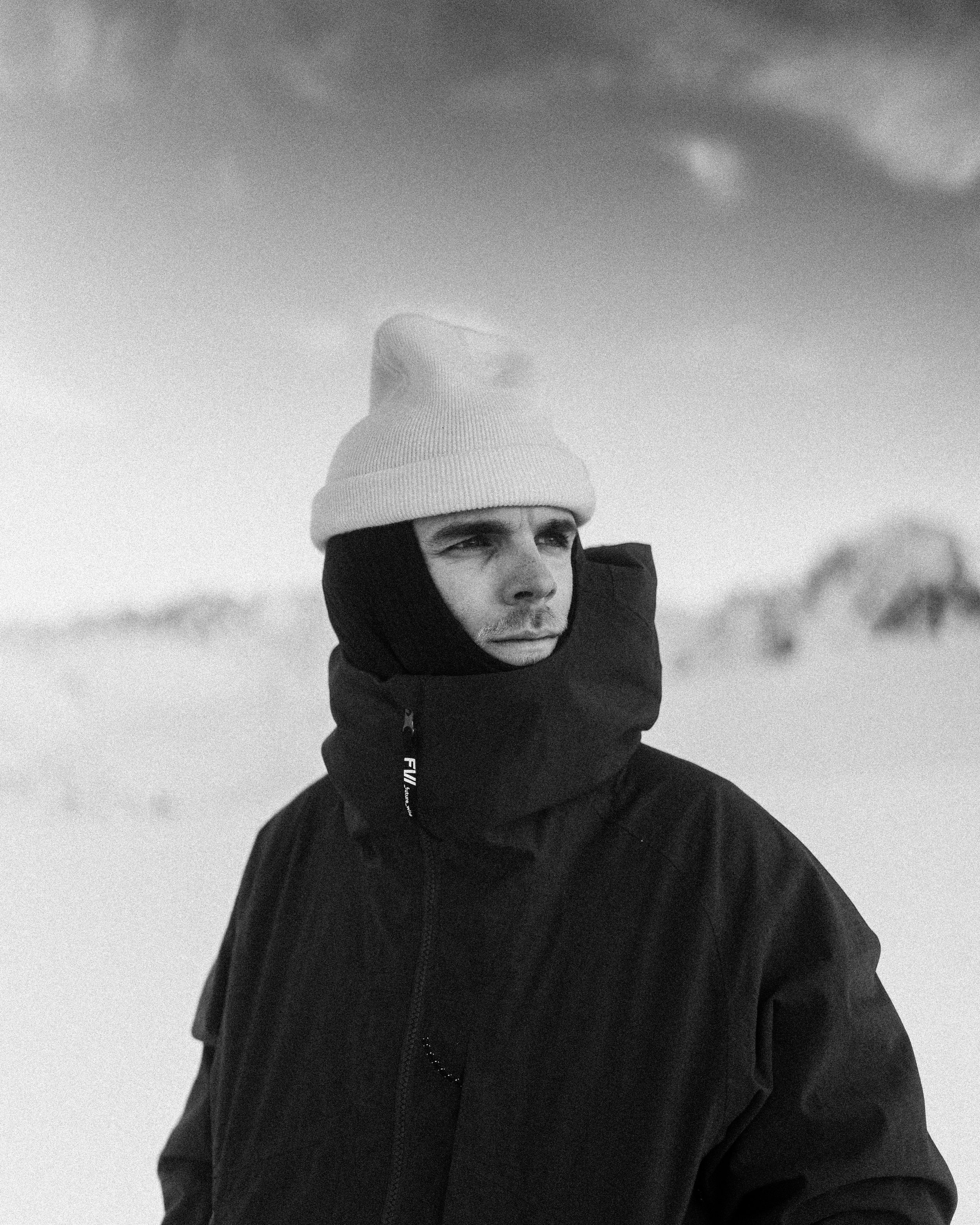 Un jeune homme portant une cagoule noire, un bonnet blanc et un blouson noir regarde légèrement vers la droite, dans un environnement en extérieur, avec un ciel nuageux en arrière-plan.