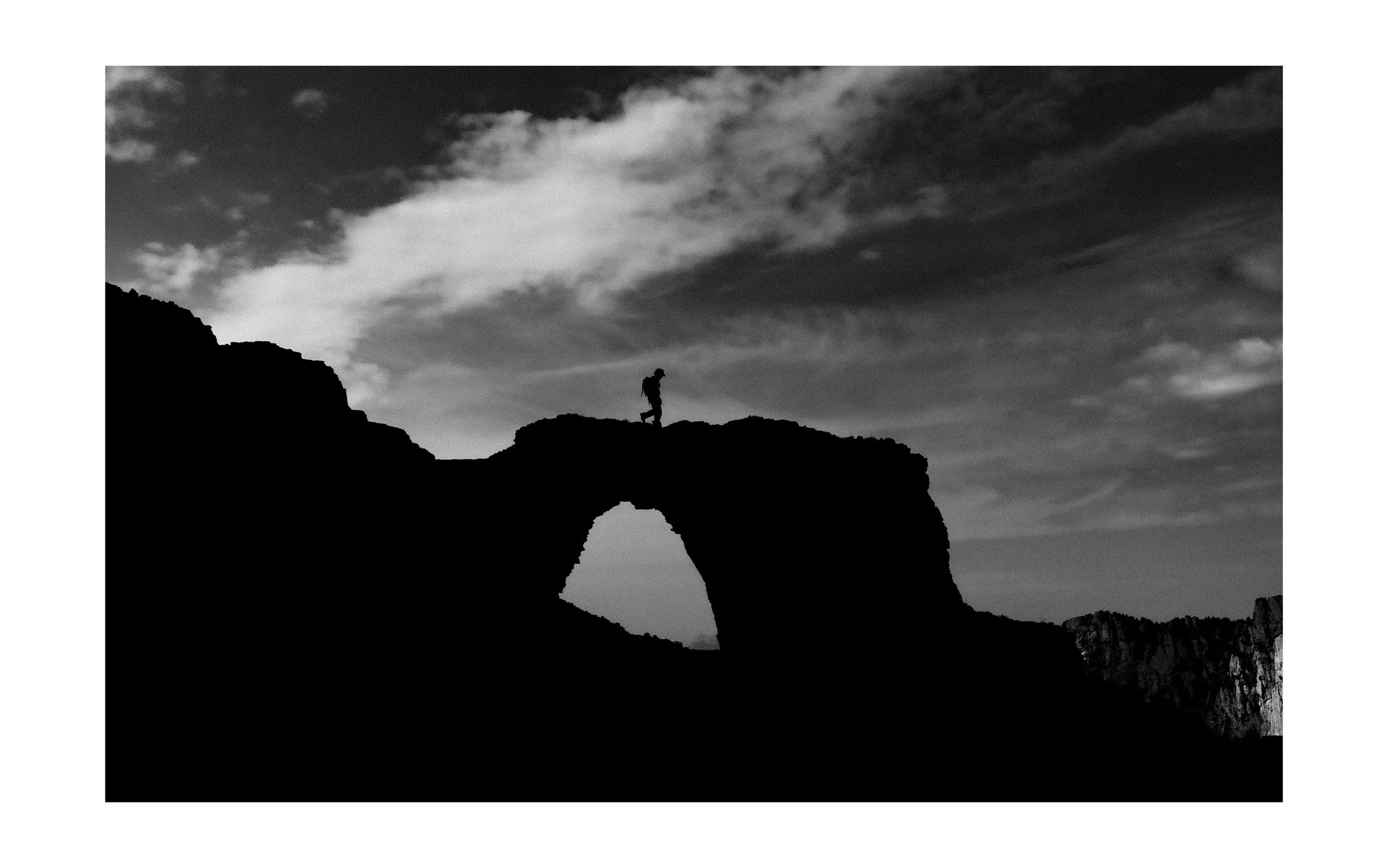 Silhouette d'une personne marchant sur un pont naturel en arc avec un ciel nuageux en arrière-plan.