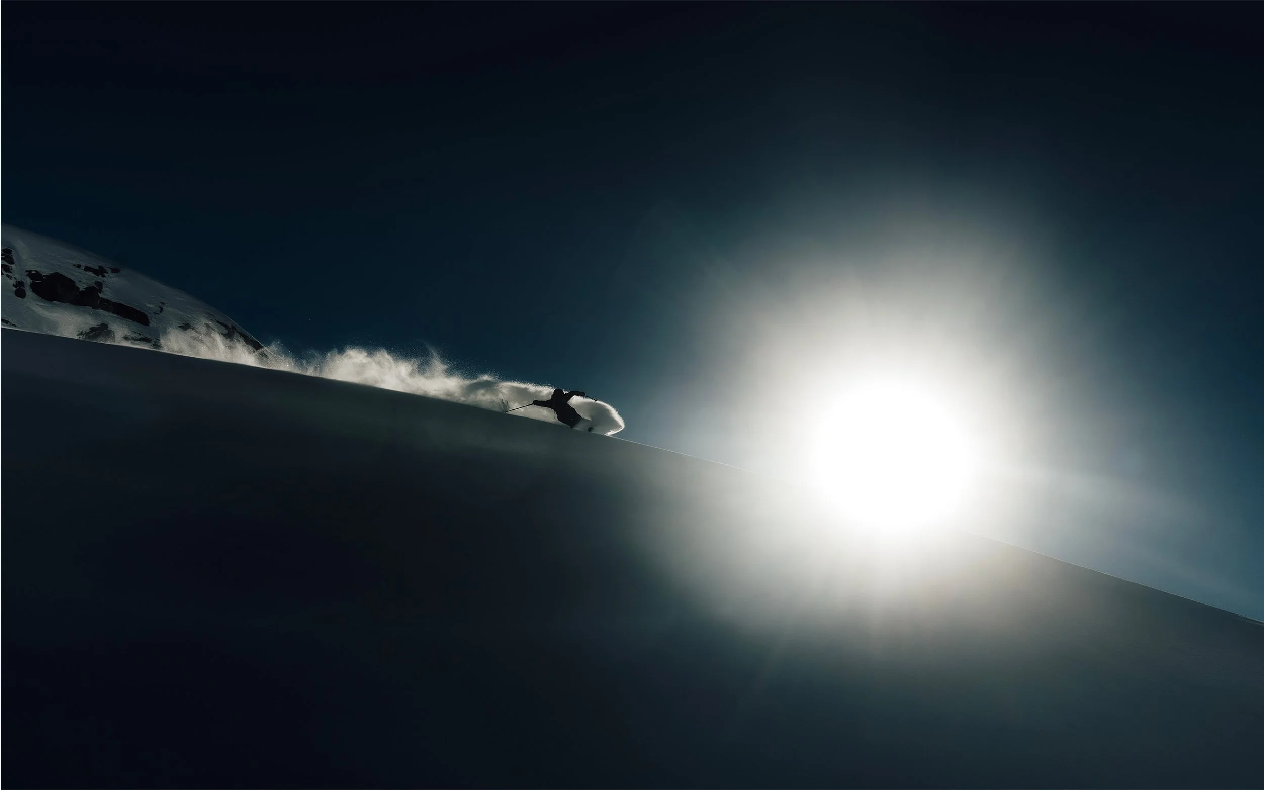 Skieur descendant une pente enneigée avec le soleil brillant en arrière-plan, créant un contraste lumineux dans un paysage de montagne.