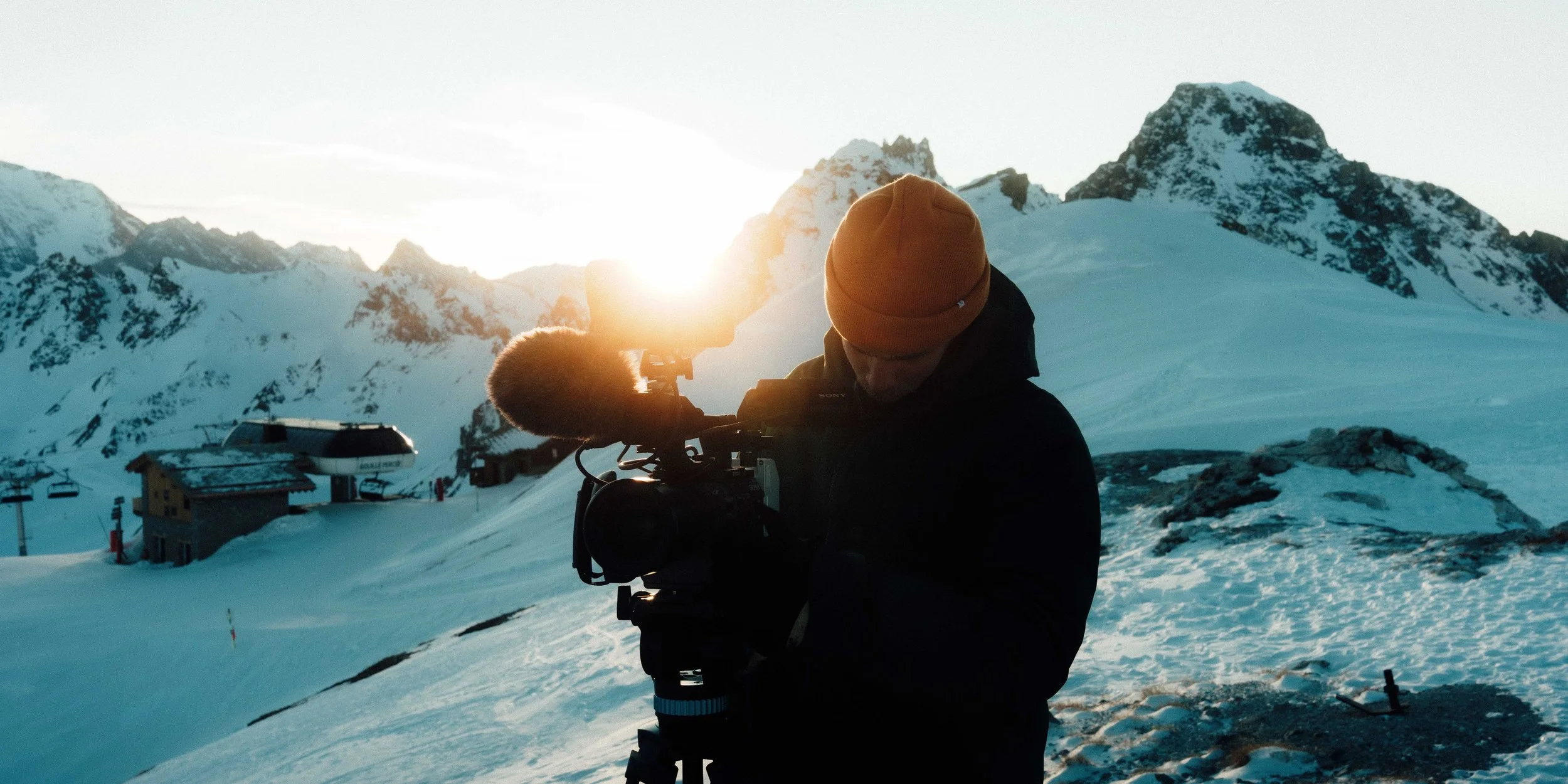 Une personne portant un bonnet orange utilise une caméra vidéo dans un paysage de montagne enneigée au coucher du soleil.