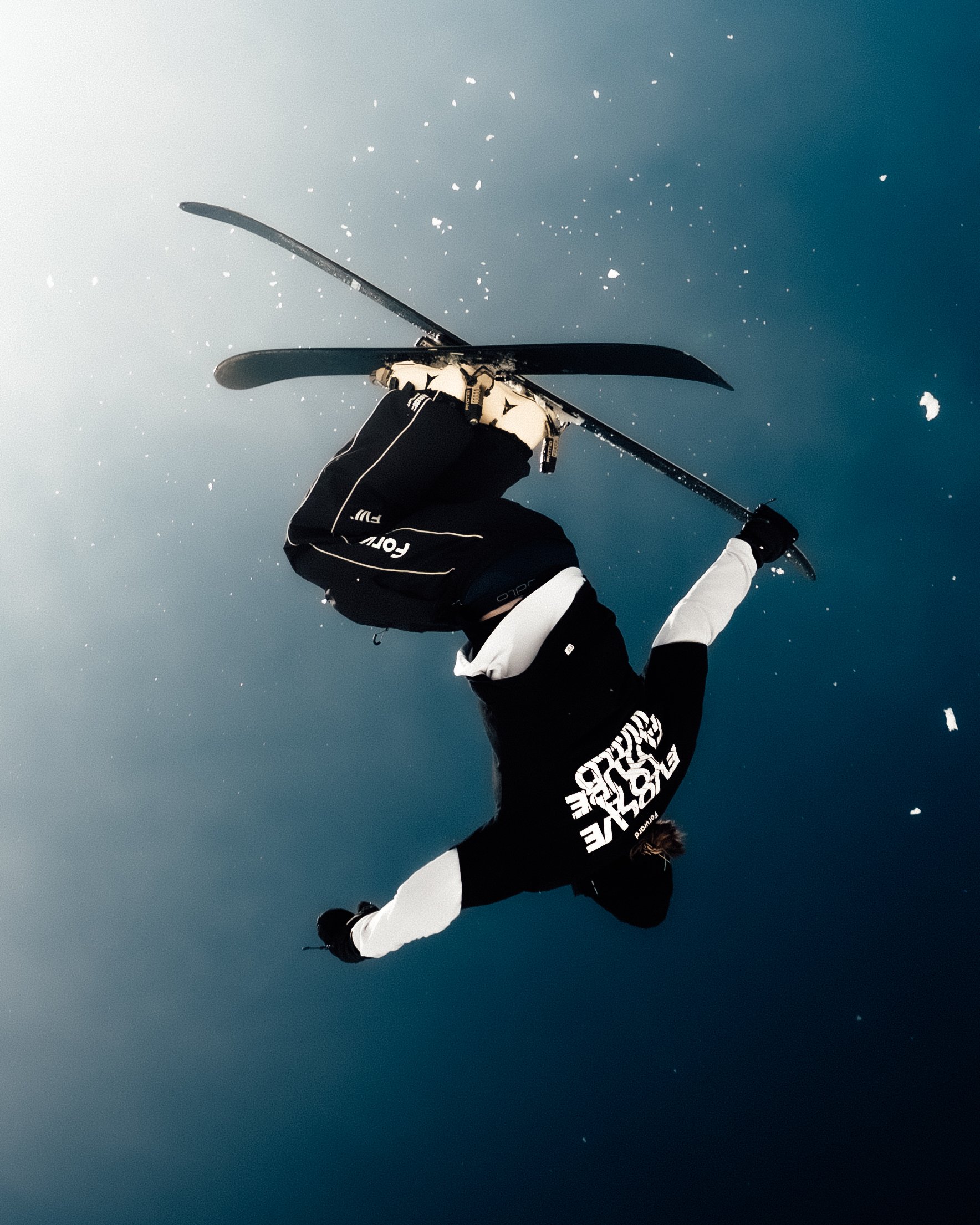 Skieur effectuant un saut en intérieur ou dans une station de ski, portant une veste noire avec des inscriptions blanches, casque et gants, avec un fond de ciel clair et quelques petits nuages.