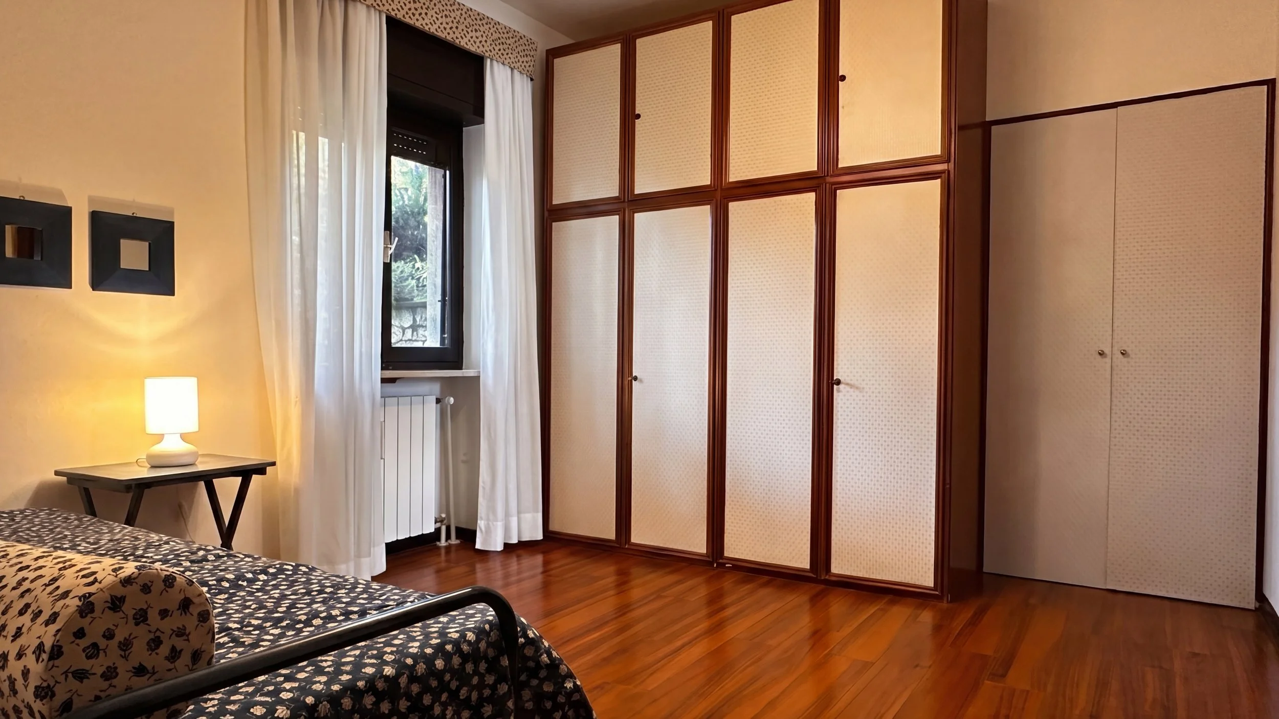 Camera da letto con armadio a muro, letto con copriletto nero con pois bianchi e due cuscini, tavolino con lampada. Finestra con tende bianche e frattale verde visibile all'esterno.