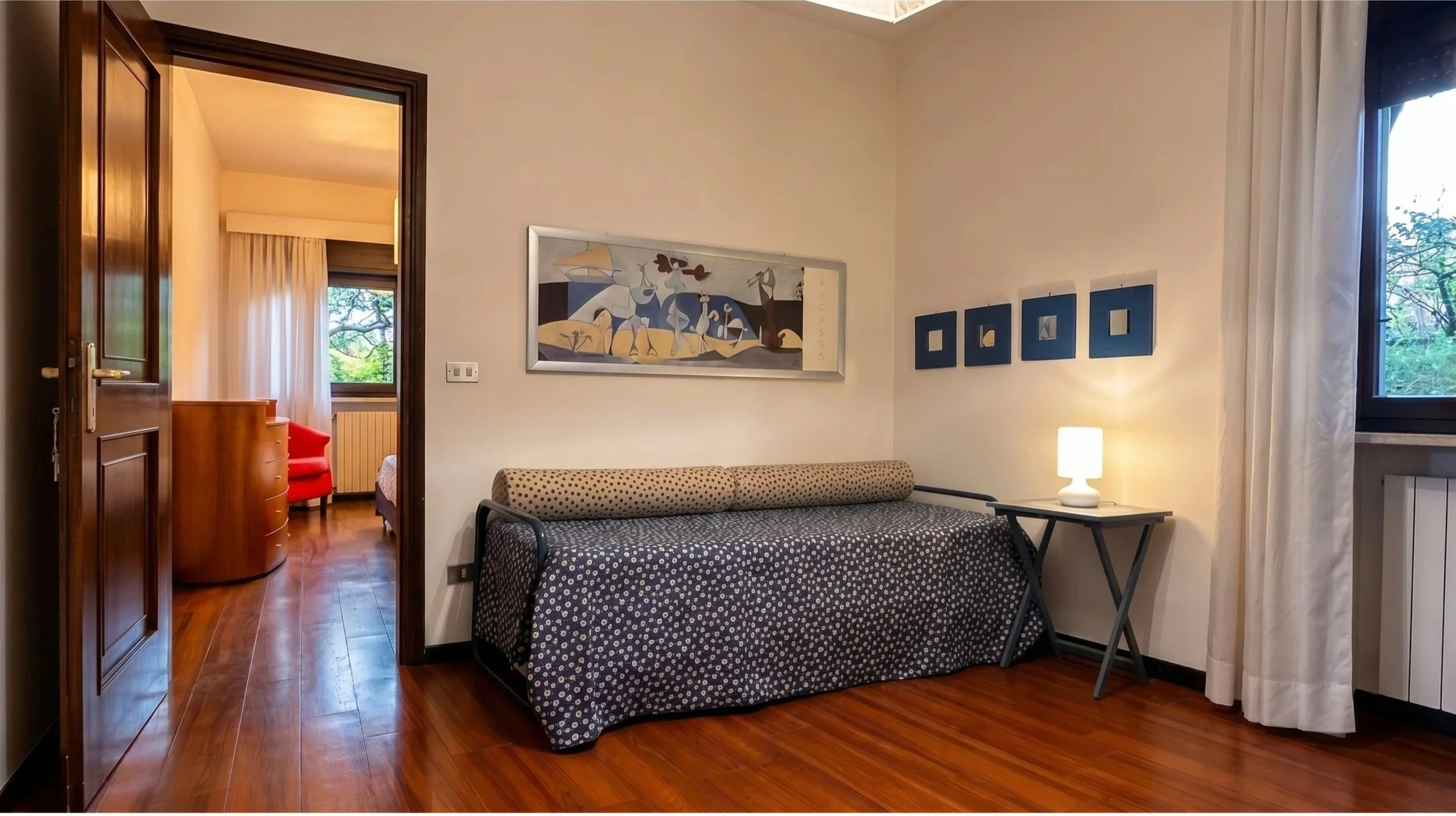 Soggiorno con divano, quadro e lampada, apertura su camera con letto, armadio, poltrona rossa e vista su giardino.