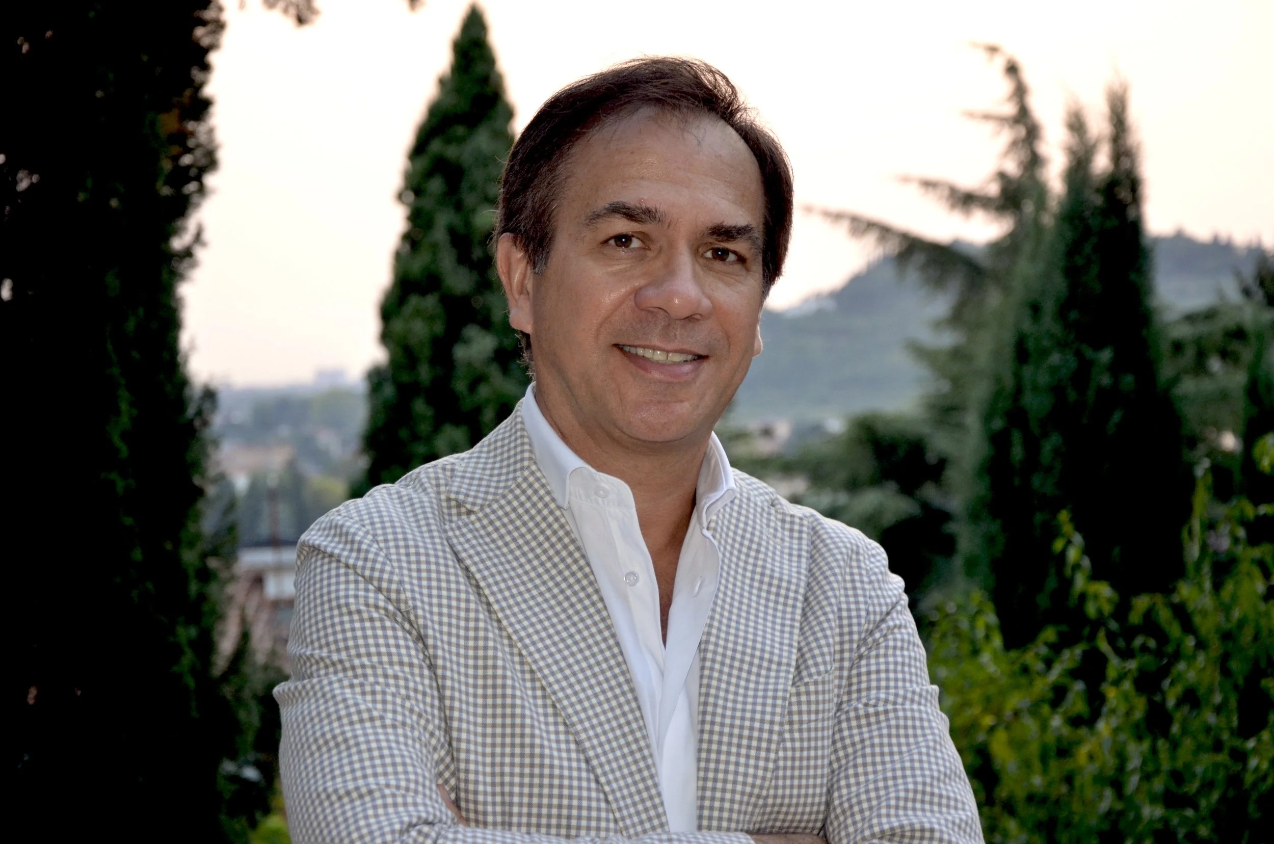 Uomo sorridente con giacca a quadri e camicia bianca si trova all'aperto, con sfondo alberi e cielo durante il tramonto.