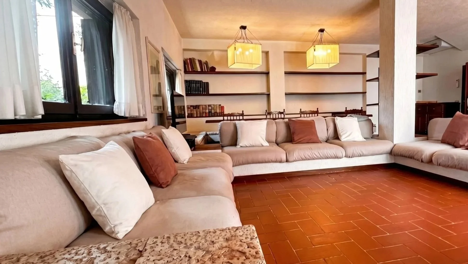 Interno di un soggiorno con divano beige, cuscini bianchi e terracotta, scaffali alle pareti e due lampadari gialli.