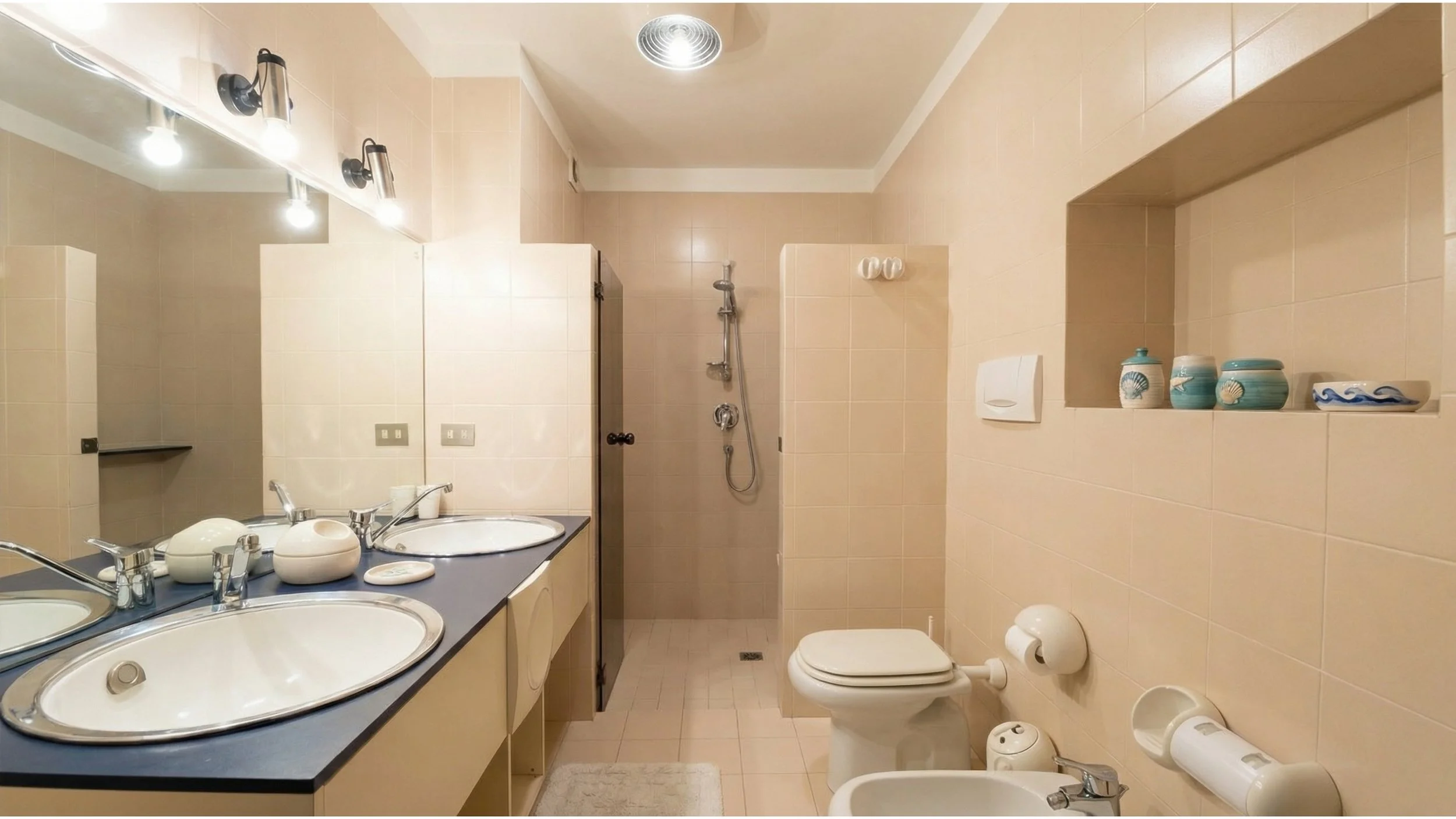 Bagno padronale con due lavabi, un WC, un bidet, un'ampia doccia , pareti rivestite di piastrelle beige, e specchio panoramico a tutta parete.