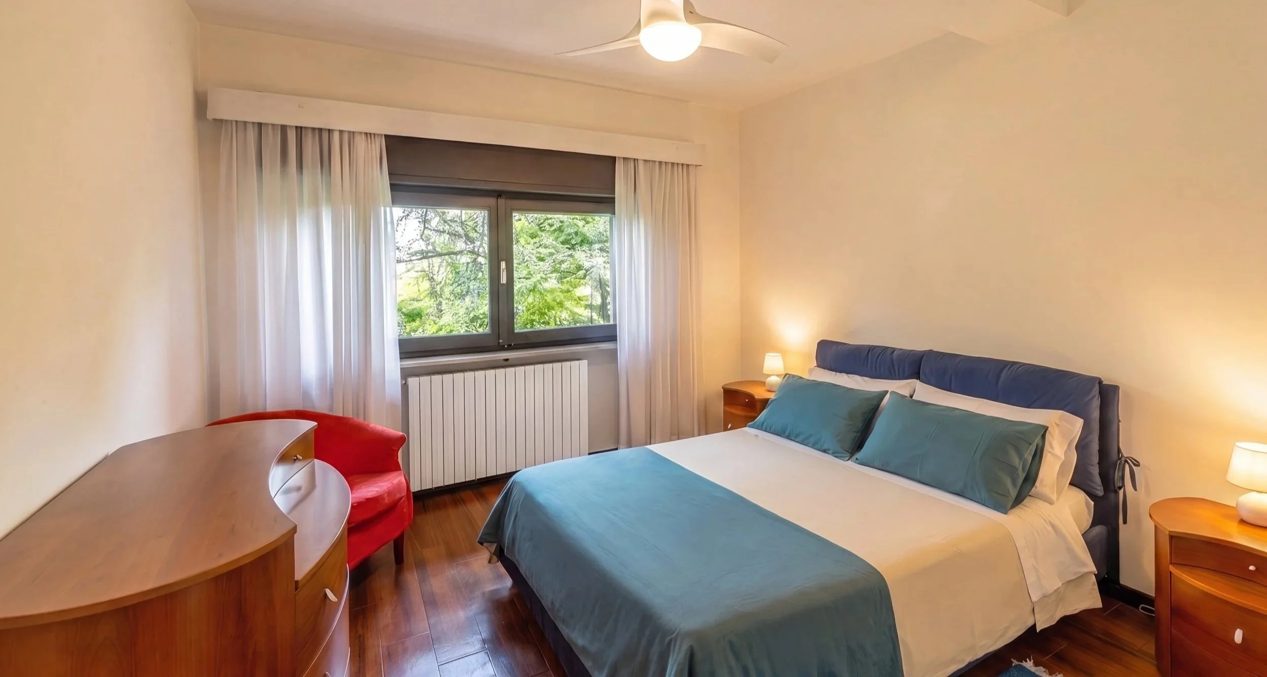 Camera da letto con letto matrimoniale, cuscini verdi, due comodini con lampade, finestra con tende bianche e vista su alberi, scrivania con sedia rossa, pavimento di legno.