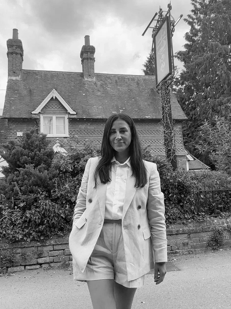 A woman standing outdoors in front of a brick house with pitched roof and chimney, wearing a light-colored blazer and shorts, with trees and bushes around her.