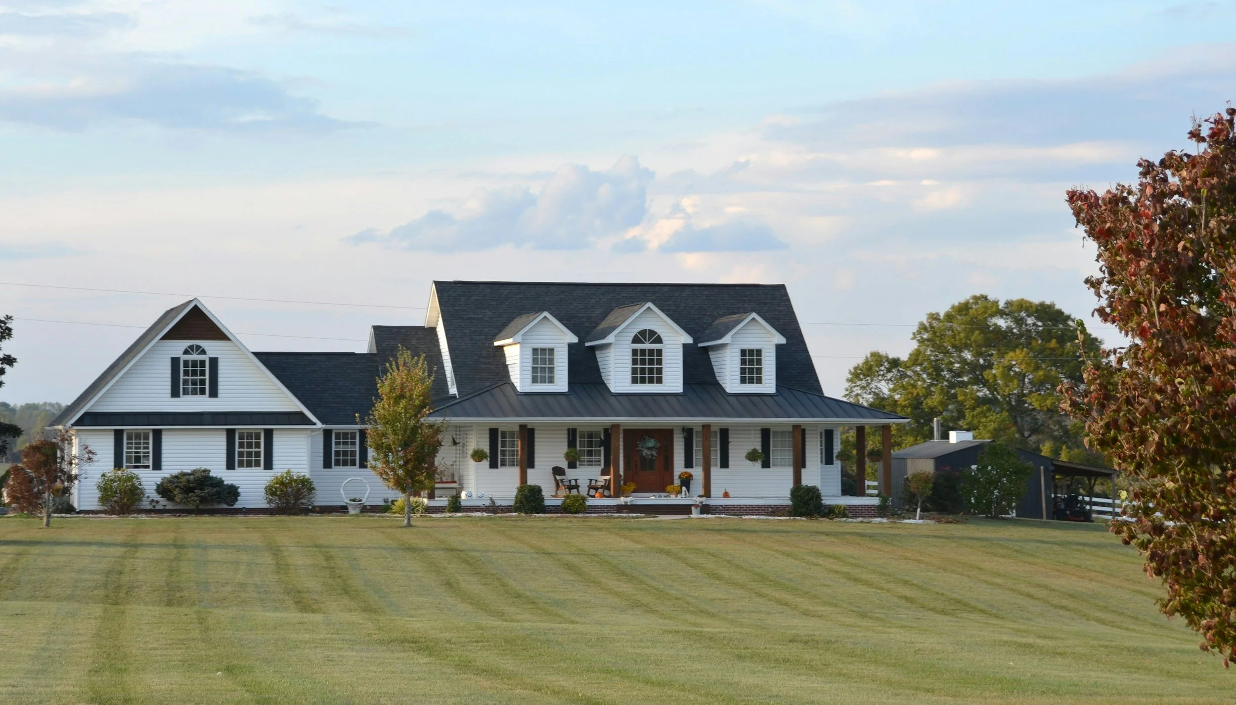 Large white farmhouse with black roof, multiple dormer windows, front porch, surrounded by green lawn and trees, clear sky overhead.