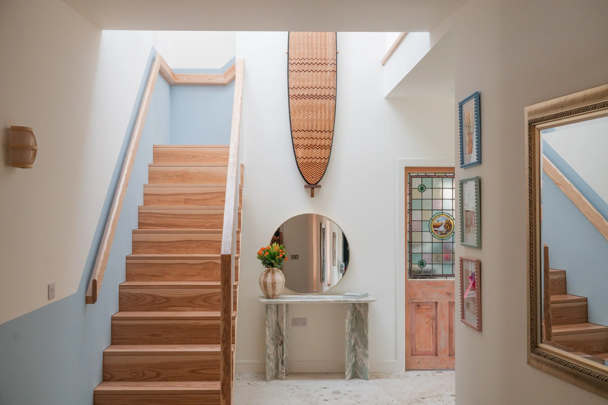 Interior view of a home with a staircase, decorative wall art, a mirror, a stained glass window, and a marble console table with a flower arrangement.