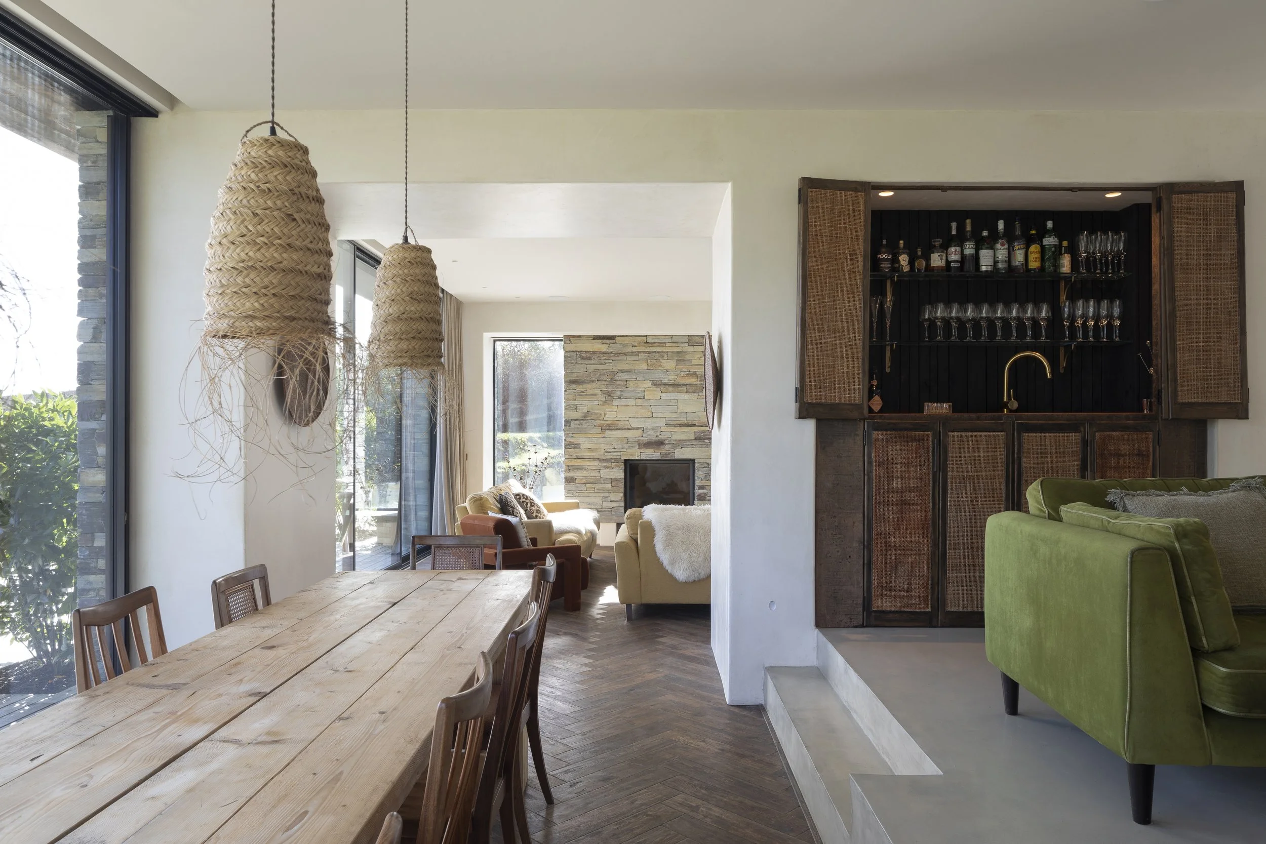 Interior view of a modern living and dining area with large glass windows, hanging woven pendant lights, a wooden dining table with chairs, a stone fireplace in the background, and a bar area with shelves of bottles and glassware.
