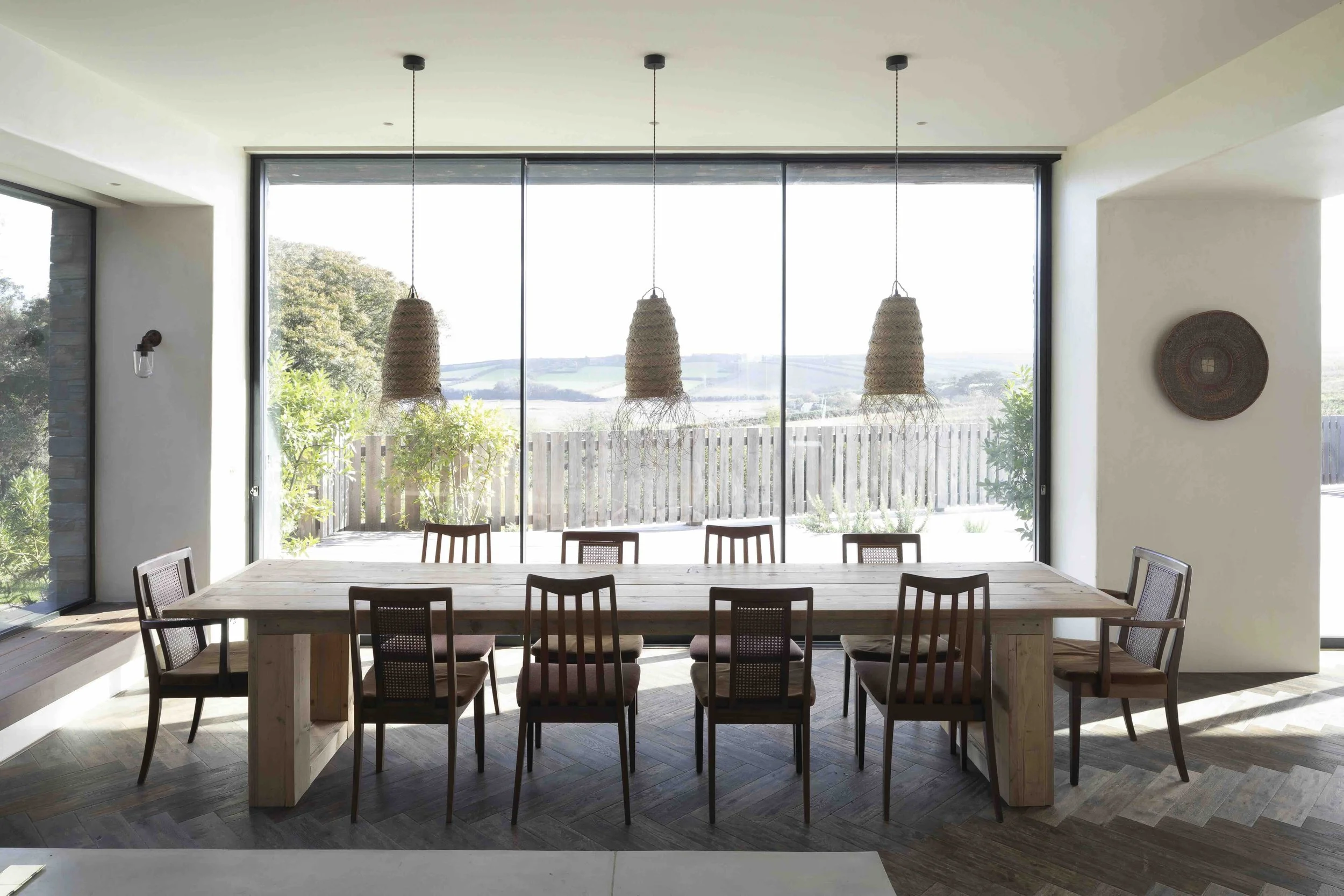 A dining area with a large wooden table and eight chairs, set in front of floor-to-ceiling windows that overlook a backyard with trees and a fence, with three pendant lights hanging from the ceiling.