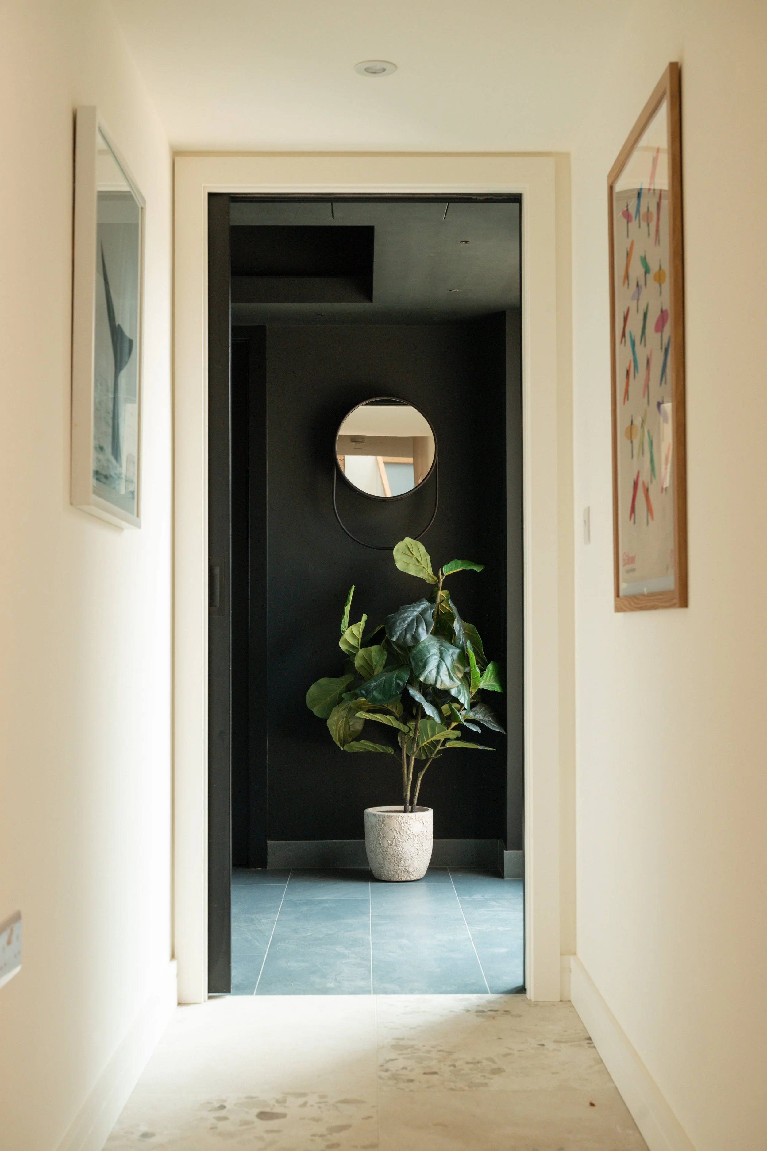 View of a hallway with cream-colored walls, a potted plant, and framed artwork on the walls. The hallway opens into a black door with a circular mirror mounted on the wall behind it. The floor has a light-colored stone or tile surface.