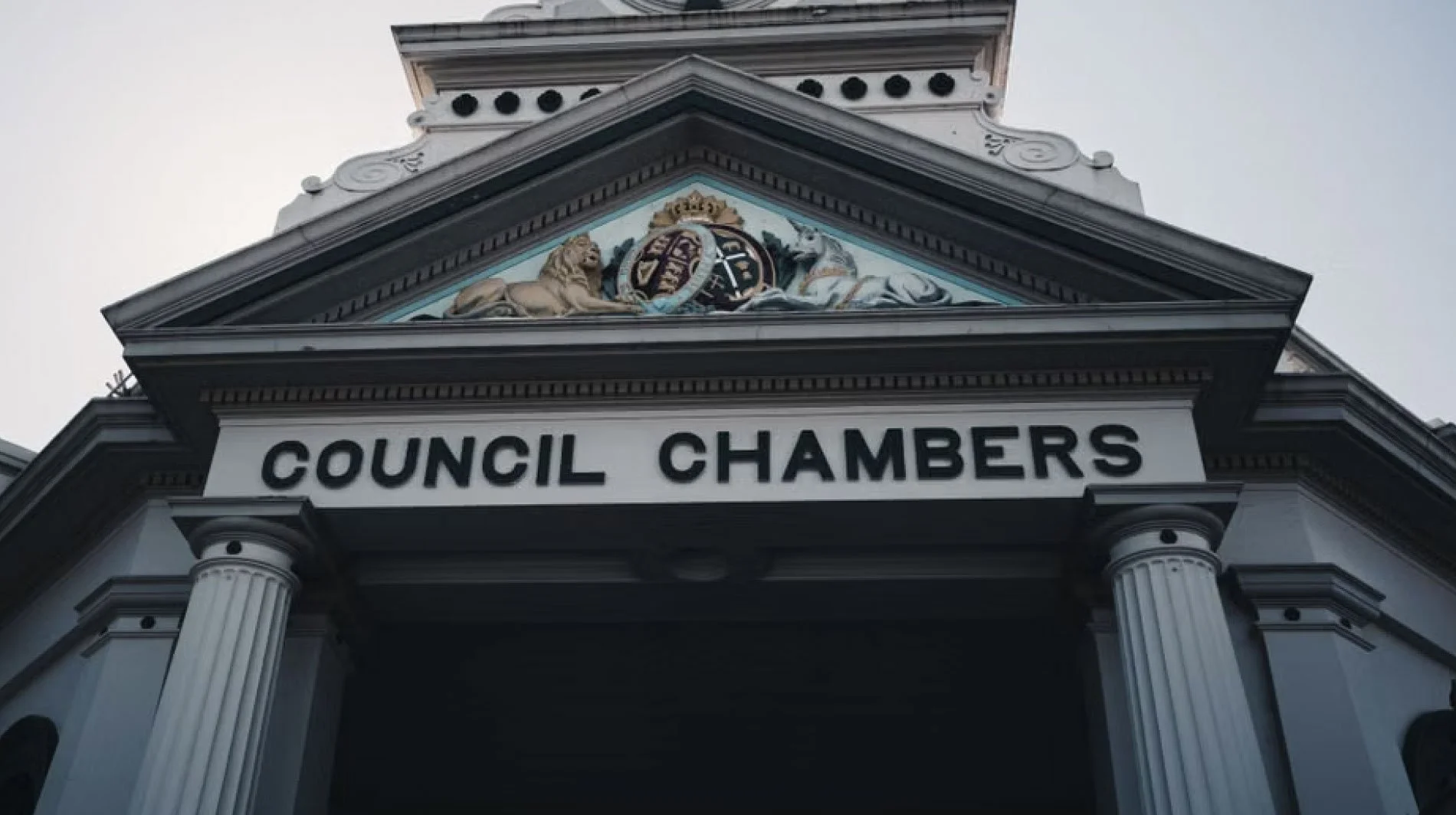 Facade of a building with a sign reading 'COUNCIL CHAMBERS', featuring classical architectural elements including columns, ornate trim, and a crest with a lion and other symbols above the sign.
