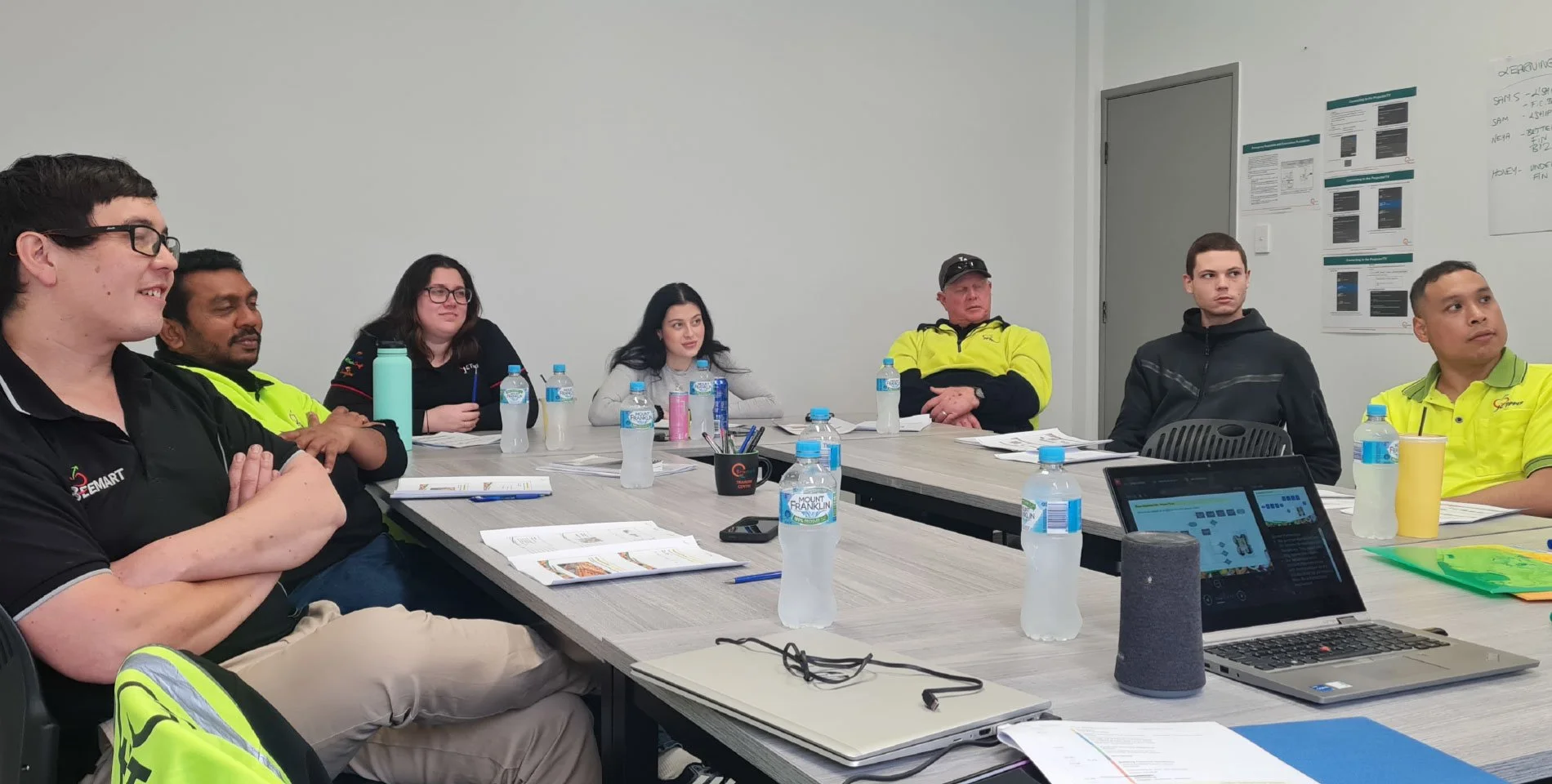 Group of seven people sitting around a conference table in a meeting room. The table has water bottles, notebooks, pens, and electronic devices. Some people are listening, others are looking at a presentation on a laptop.