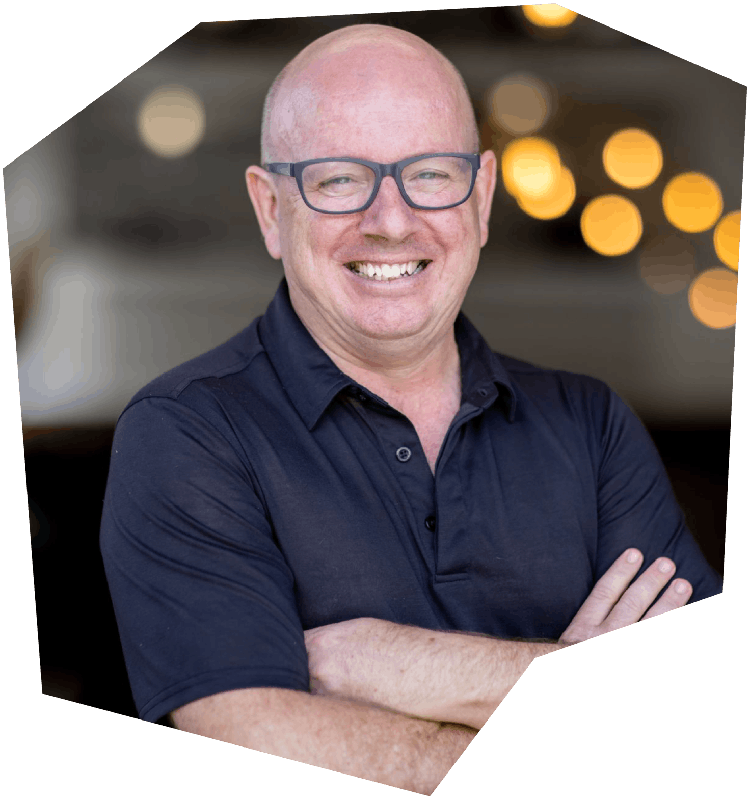 A middle-aged man with a bald head, glasses, and a big smile standing with arms crossed, wearing a dark blue shirt, in a warmly lit indoor setting.