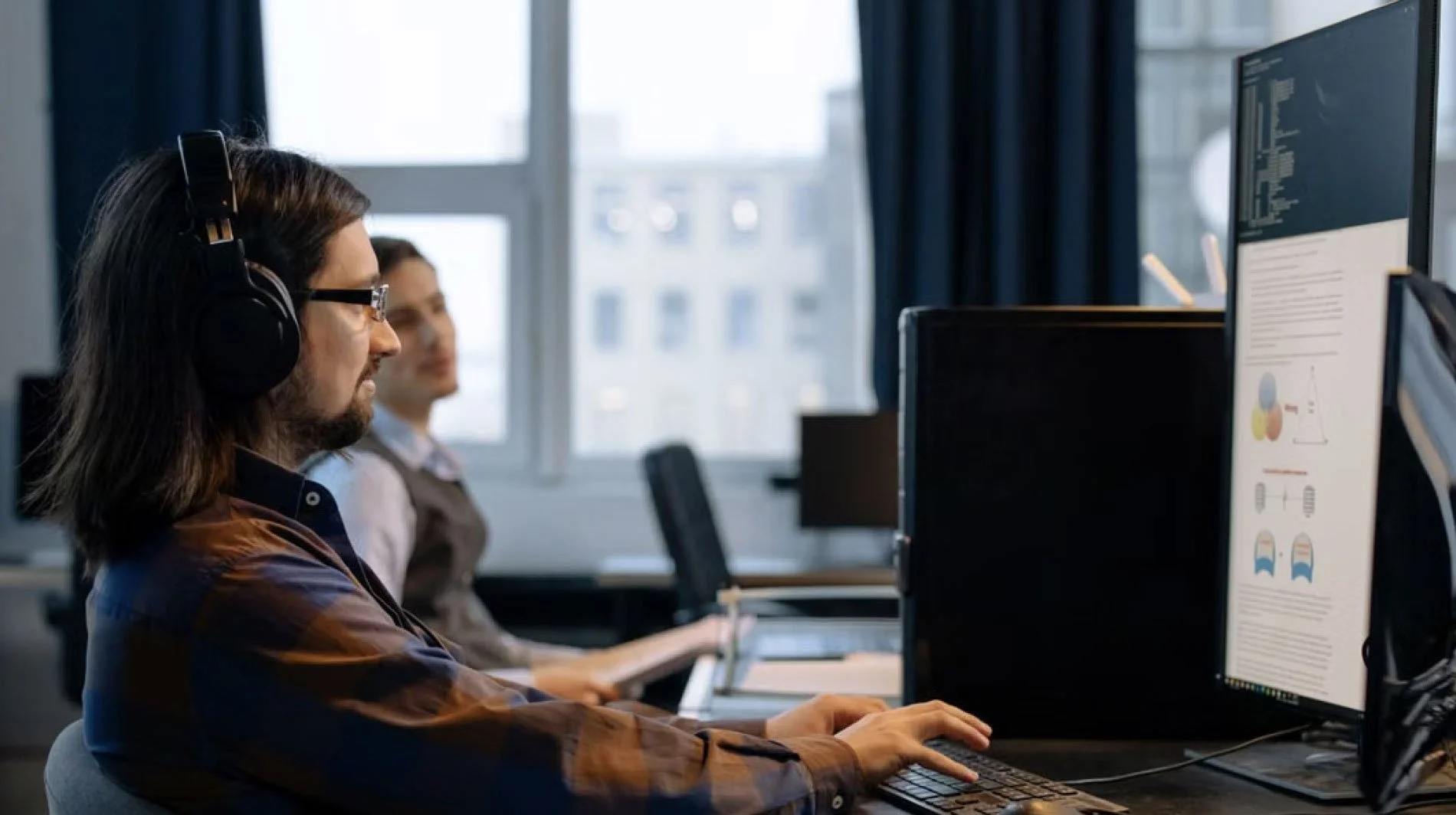 A man with long hair and glasses working at a computer with a large monitor in an office setting, with a woman in the background also working at a desk near a window.