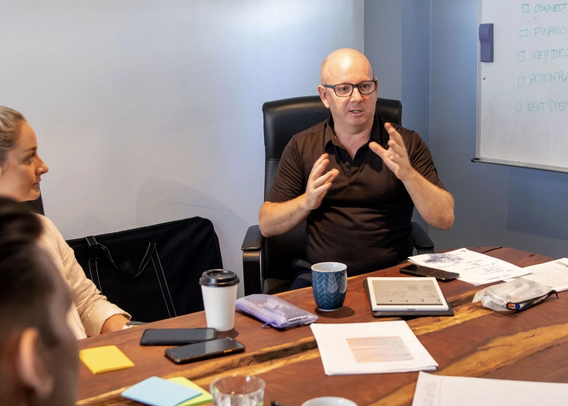 A man with glasses, bald, wearing a brown polo shirt, speaking animatedly during a business meeting in a conference room. A woman with blonde hair and a beige sweater is partially visible, sitting to his left. The table has notebooks, smartphones, a tablet, a coffee cup, and papers. A whiteboard with checkboxes and handwritten notes is on the wall in the background.