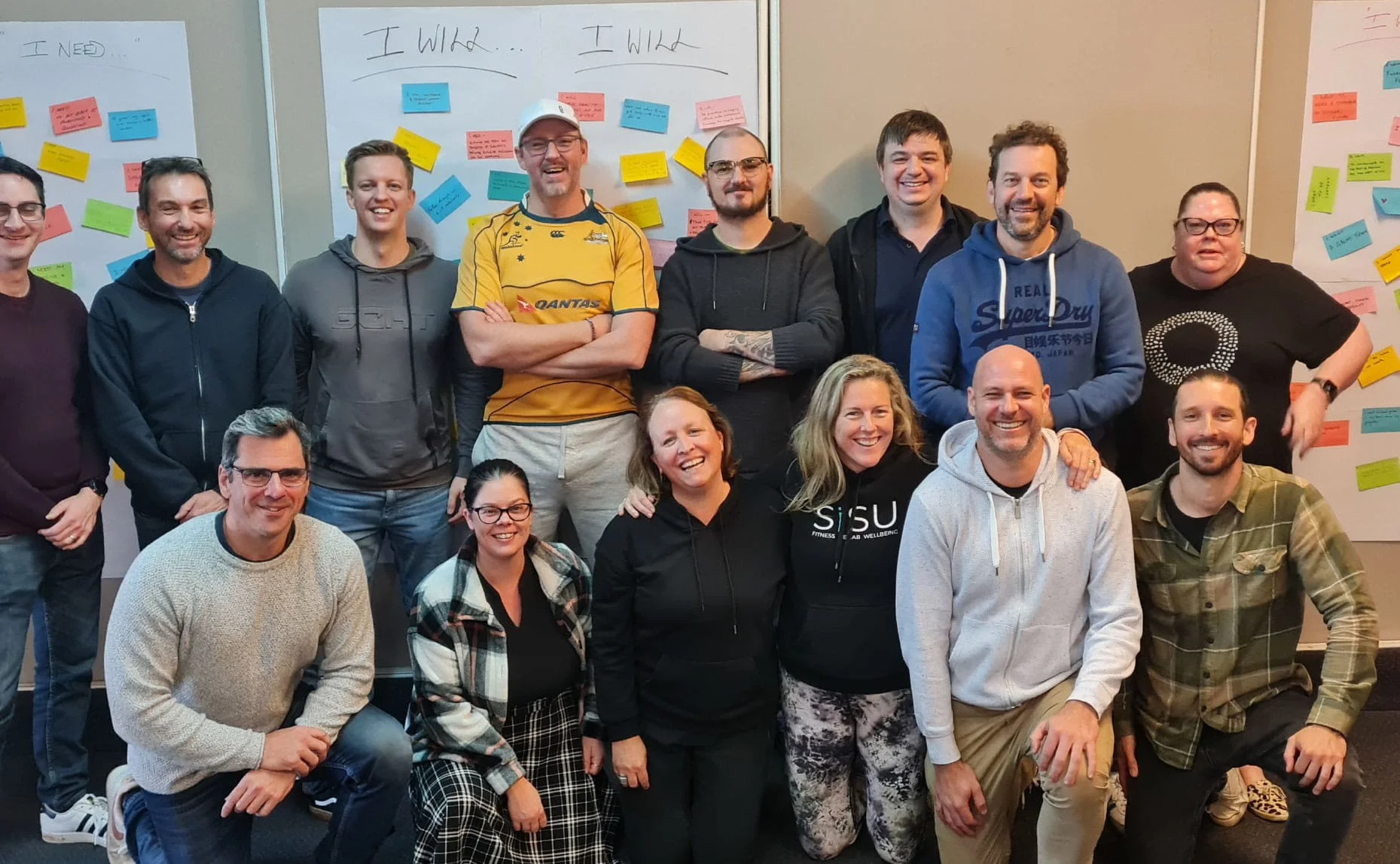 A group photo of 14 people, both men and women, smiling and posing together indoors, in front of whiteboards with colorful sticky notes and handwritten notes.