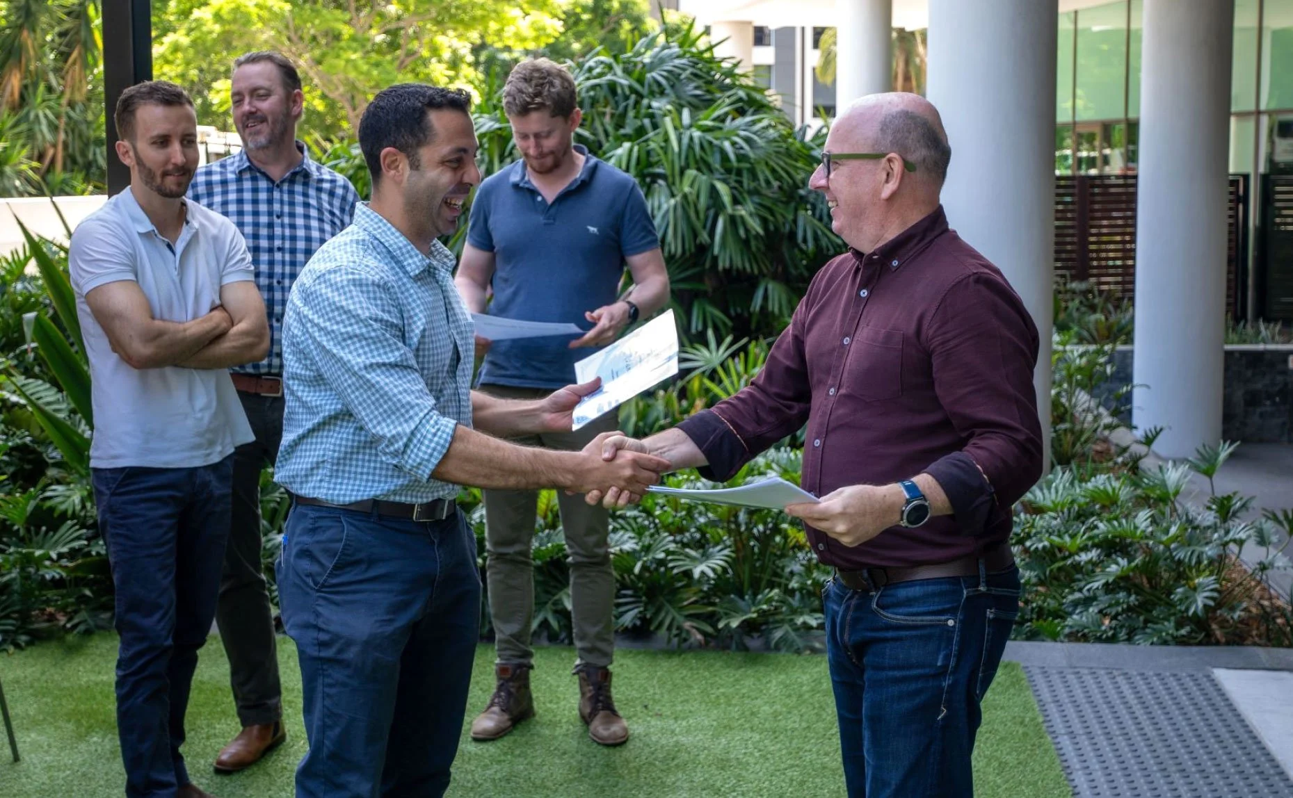A group of five men outdoors, two men shaking hands and exchanging documents, others observing and smiling.
