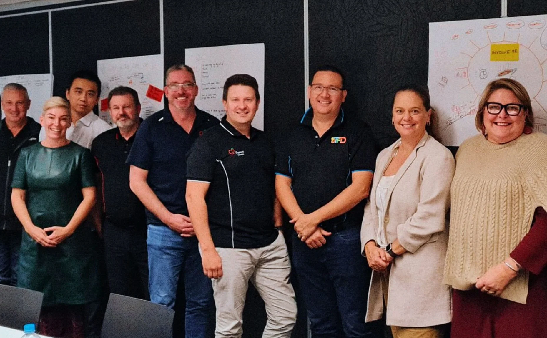 Group of nine people standing in front of a wall with charts and diagrams, smiling for the photo at a professional or conference setting.
