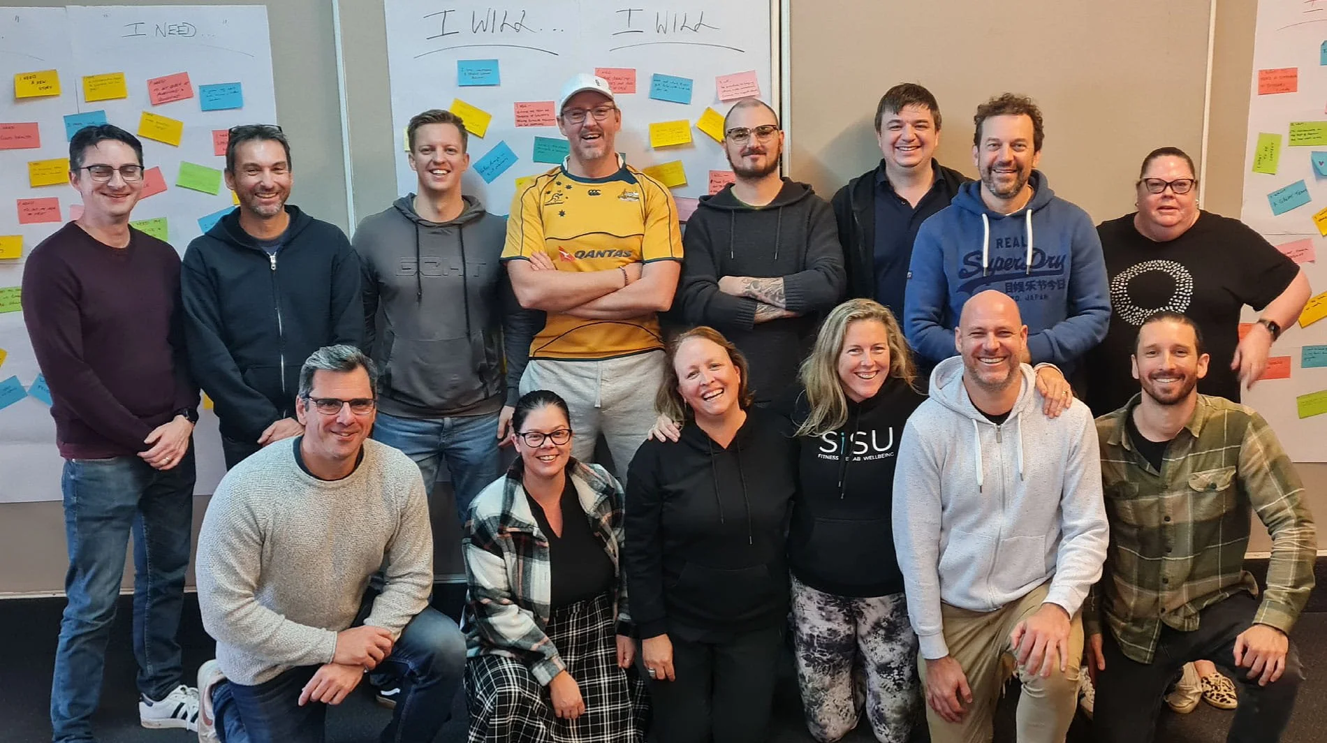 Group of about 16 people posing for a photo in front of whiteboards with colorful sticky notes, some with handwritten words like "I Need" and "I Will".