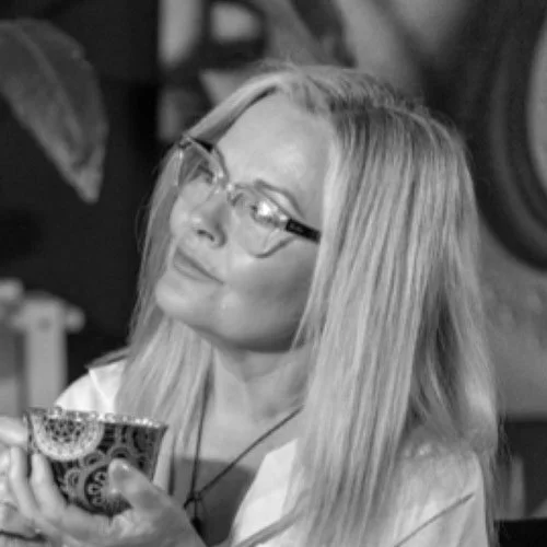 A woman with long, straight blonde hair and glasses holding a cup, looking thoughtfully.