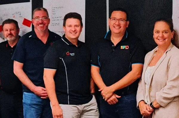 Five people standing together indoors, smiling for the photo. Four men and one woman, with whiteboards and blackboard behind them.