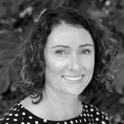 A woman with curly hair smiling, wearing a polka dot top, outdoors with leafy background.