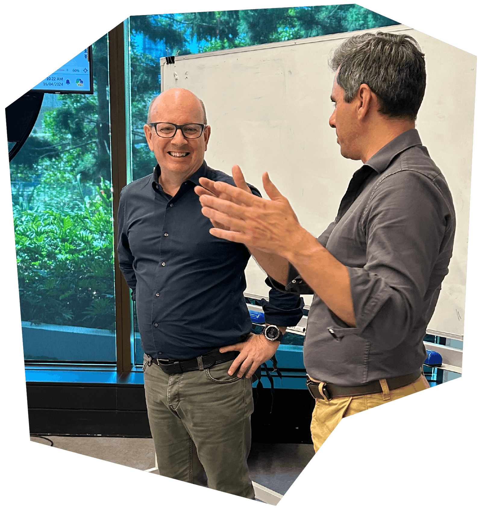 Two men having a conversation in a boardroom with a whiteboard and a monitor displaying the time and weather in the background.