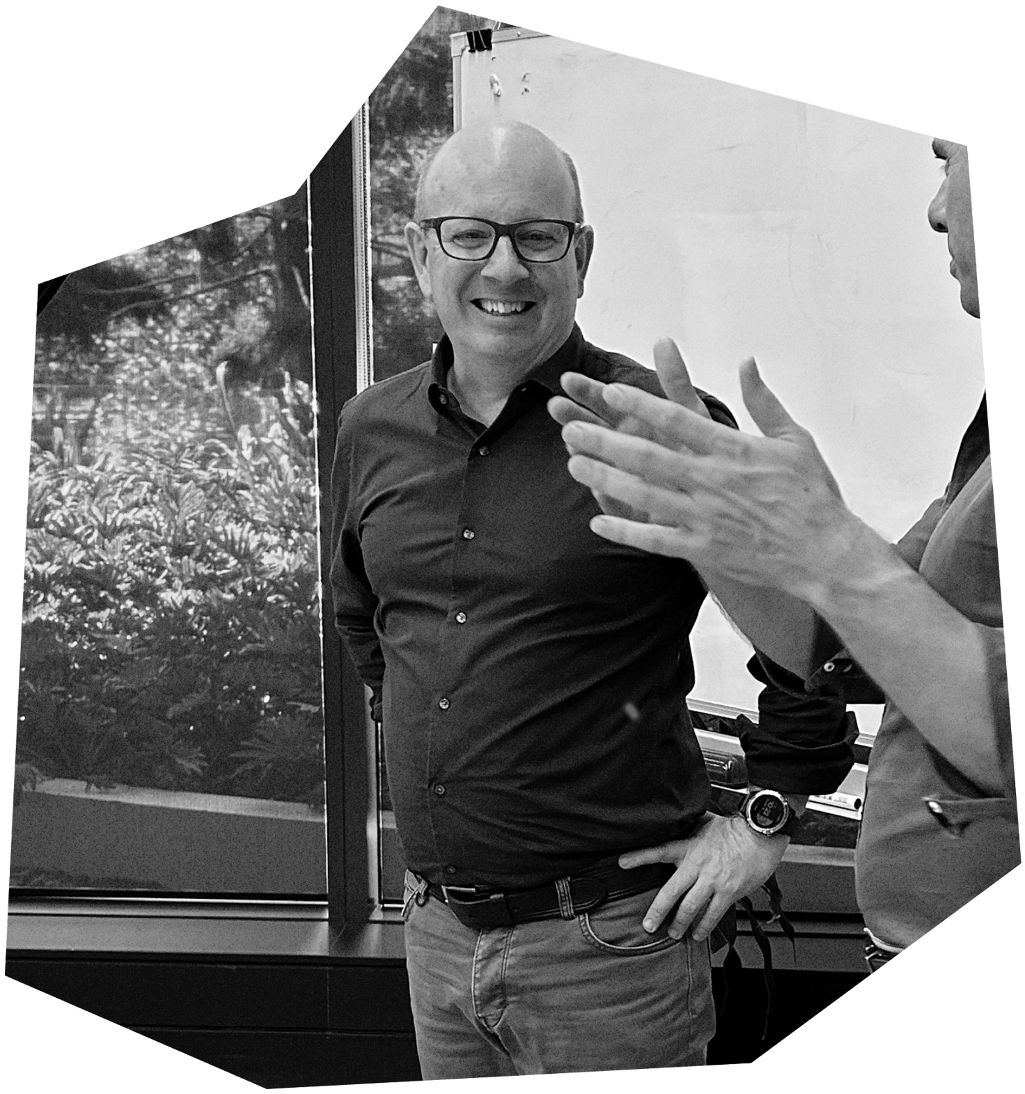 Black and white photo of two men having a conversation indoors. The man facing the camera is smiling, wearing glasses, a button-up shirt, and jeans. The other man, partially visible, is gesturing with his hand. There is a large window and a poster of plants behind them.
