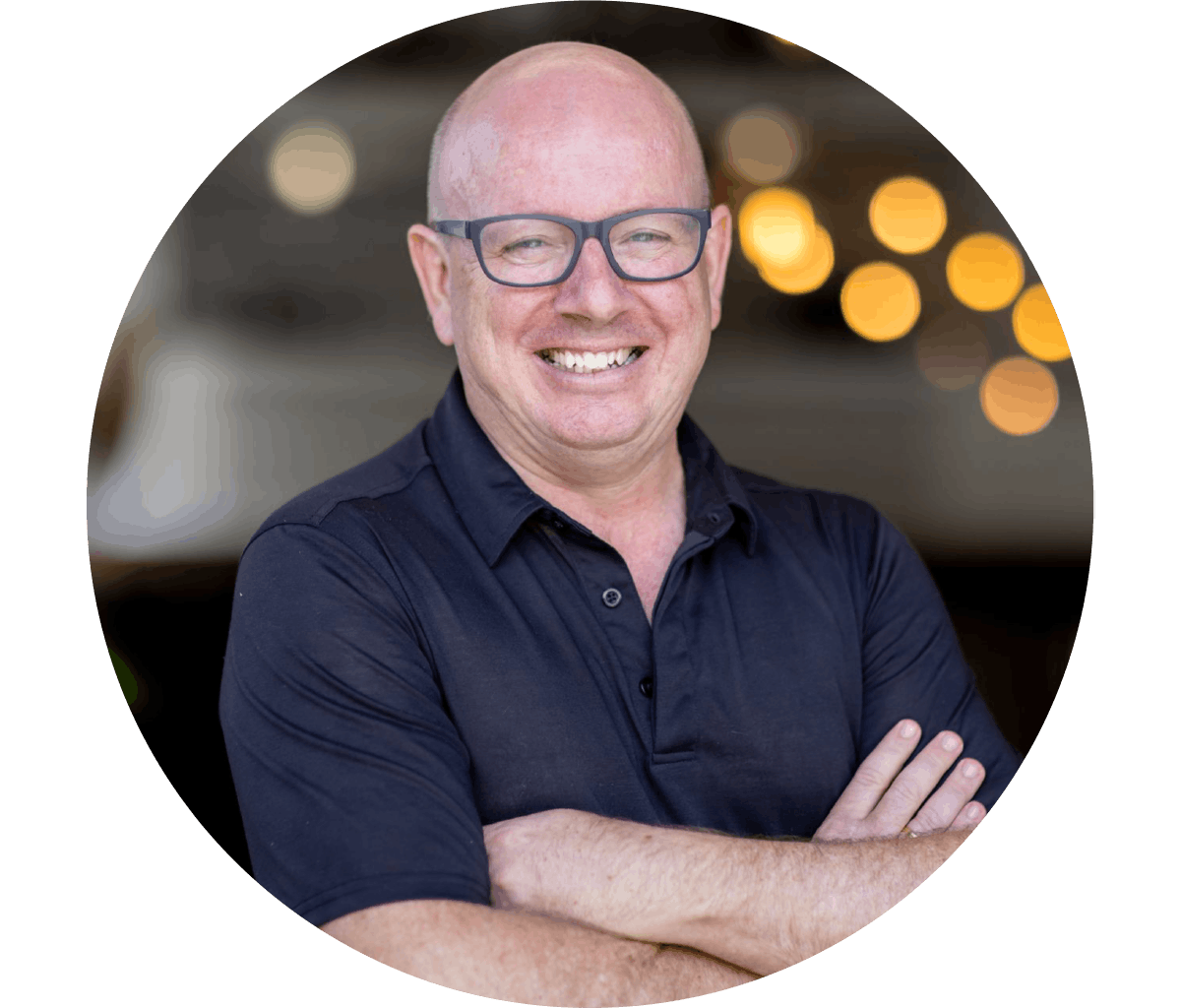 Dan Wright is smiling, with black glasses, wearing a dark button-up shirt, standing outdoors with blurred lights in the background.