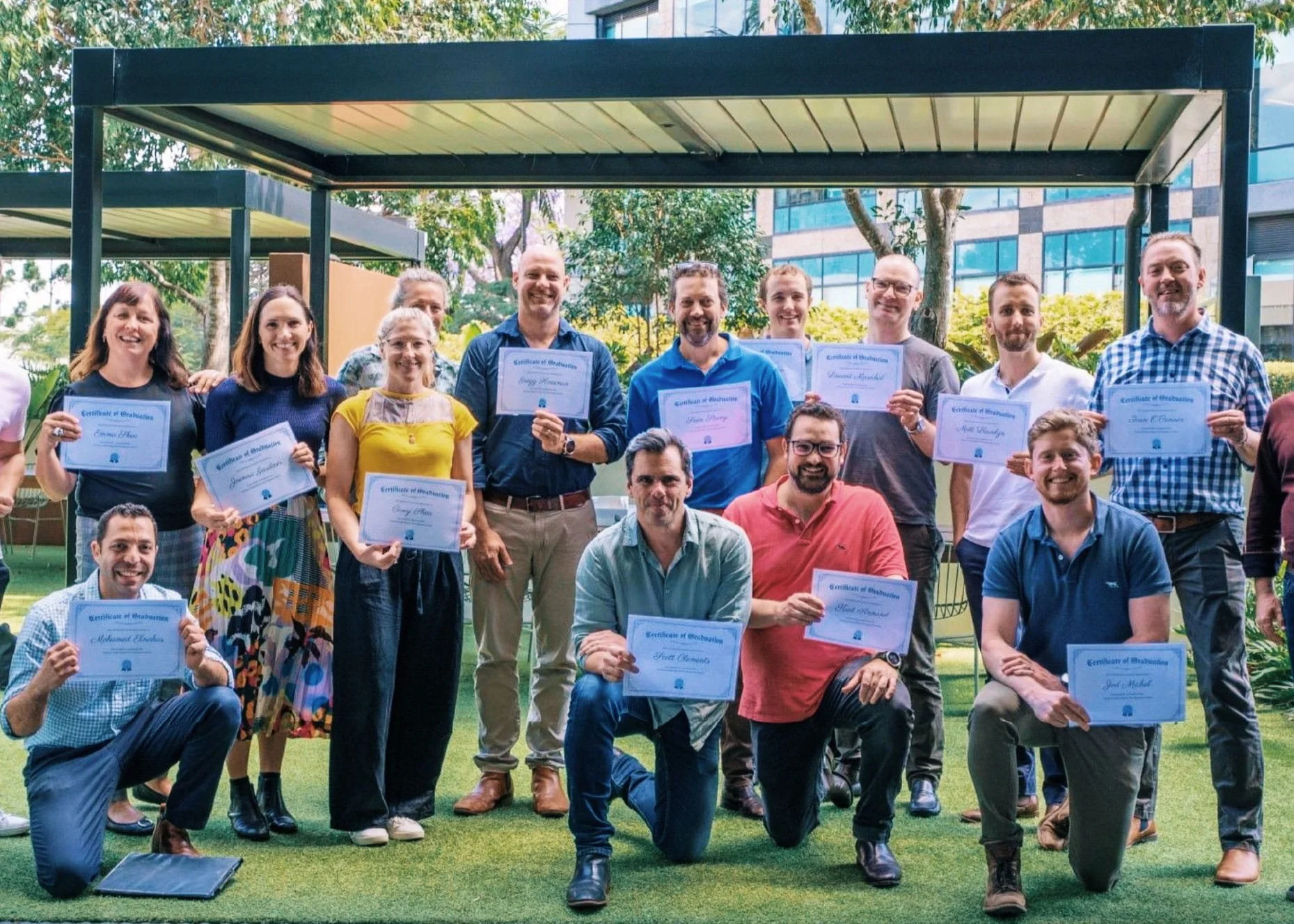 Group of people outdoors at a graduation or awards ceremony, holding certificates, with modern office buildings and trees in the background.