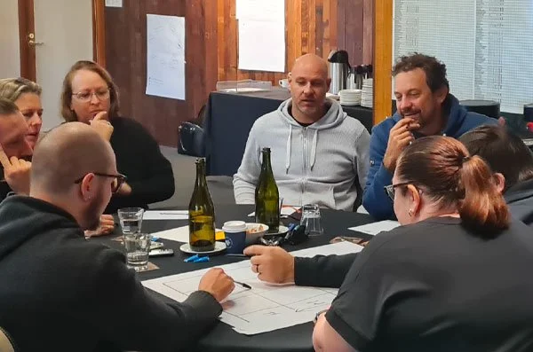 Group of seven people sitting around a table in a room, engaged in a discussion or meeting, with bottles, glasses, and papers on the table.