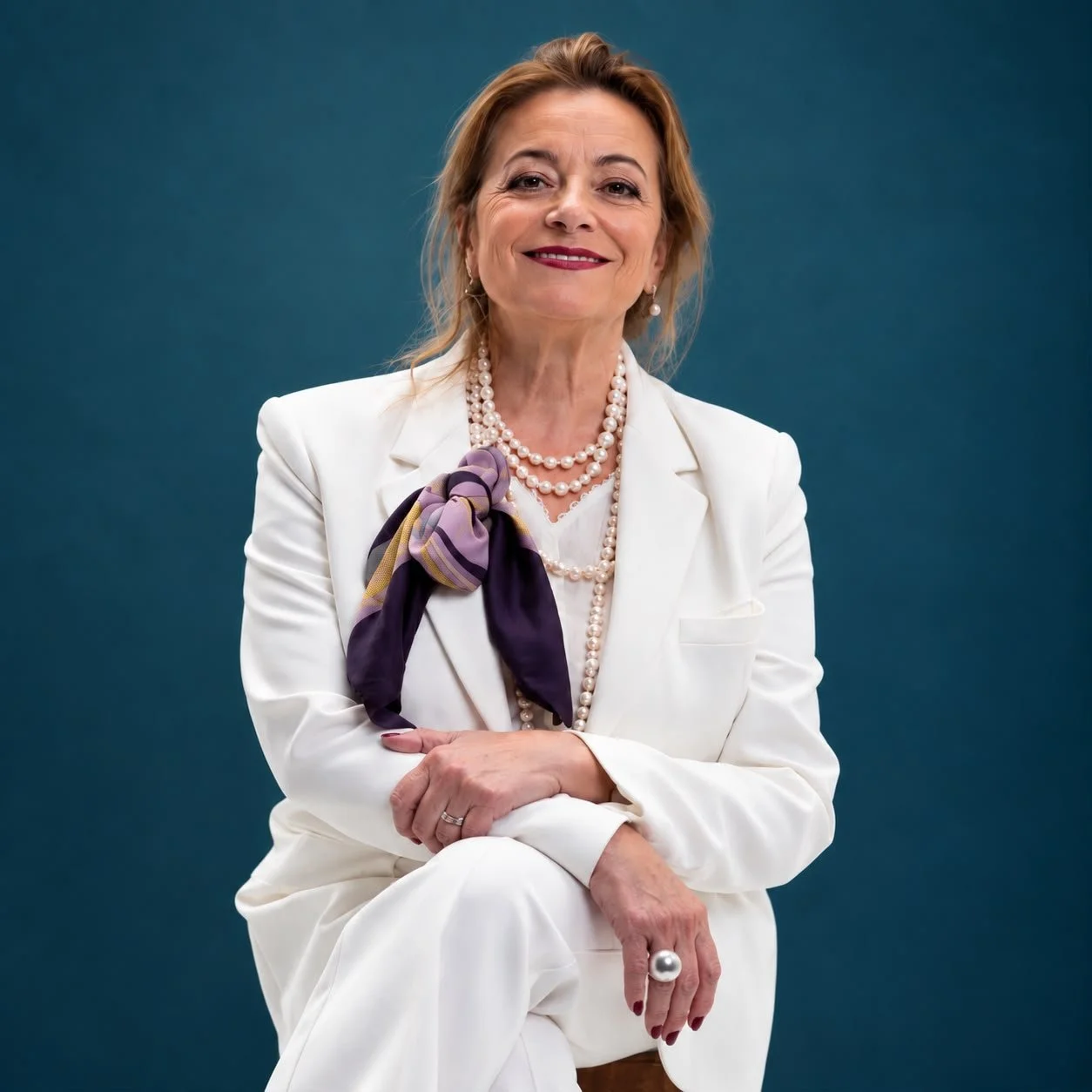 Mujer mayor con cabello rubio, vestido blanco y accesorios de perlas posando contra un fondo azul.