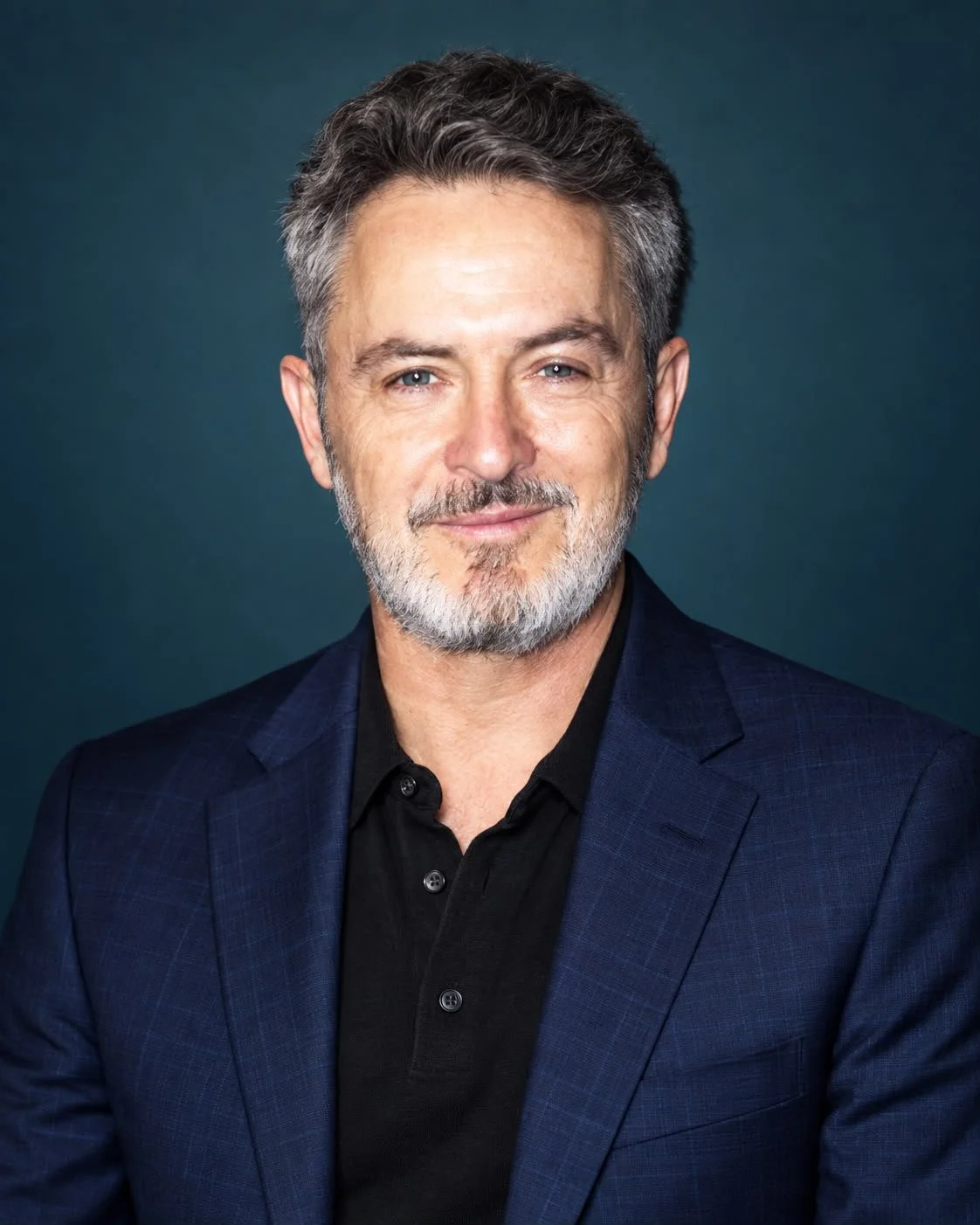 Retrato de un hombre de cabello gris y barba, vistiendo un traje azul y una camisa negra, sonriendo levemente, fondo oscuro.