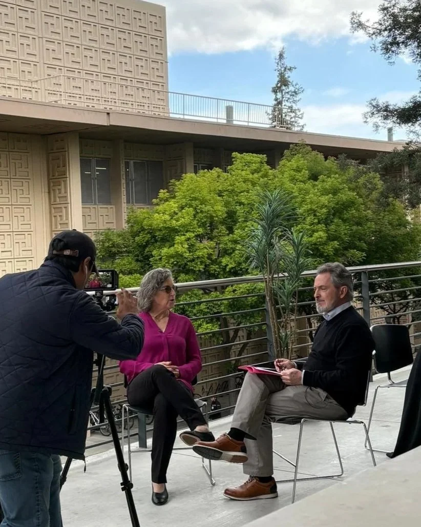Una entrevista grabada en un espacio abierto con dos personas sentadas en sillas, una mujer con blusa rosa y un hombre con camisa azul y suéter negro, mientras un camarógrafo los filma.