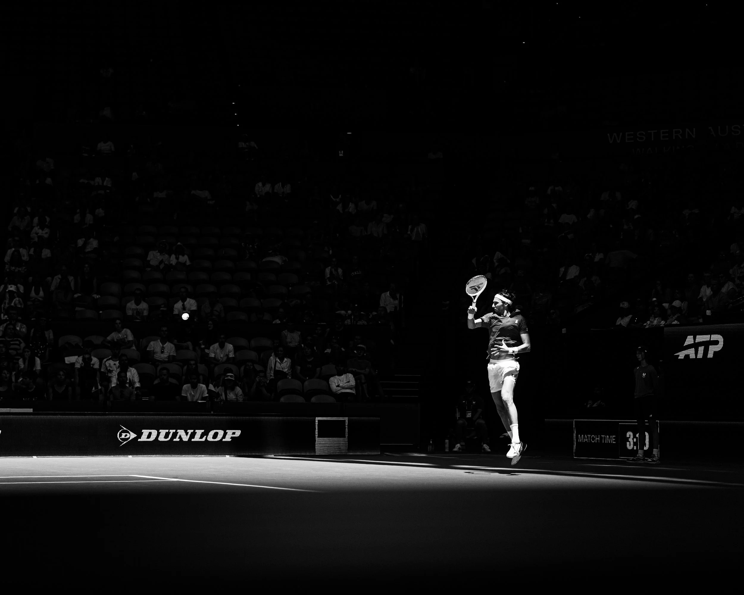A tennis player is jumping to hit a ball on a tennis court during a match, with spectators watching in the background, black and white image.