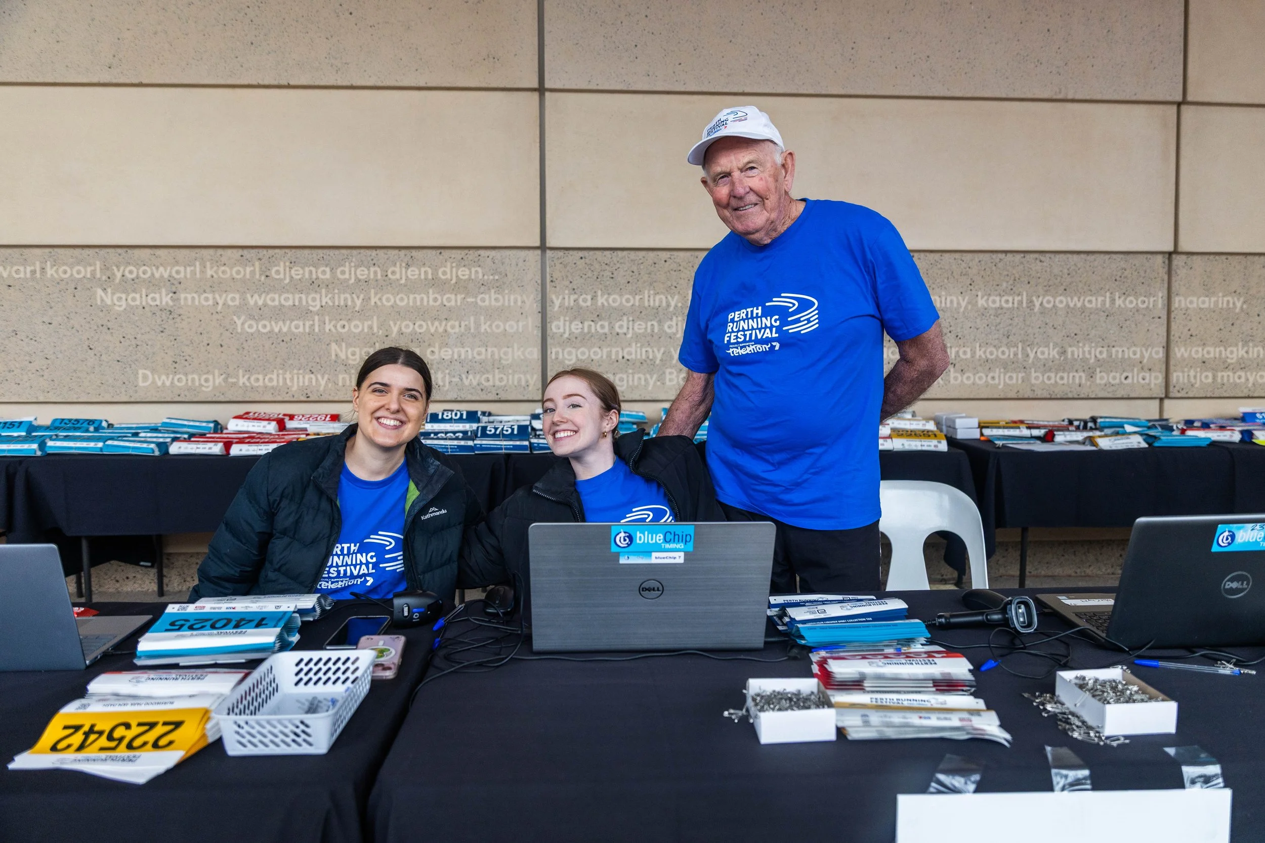 Three smiling people, two young women and one older man, sitting and standing at a table with race bibs, computers, and race timing equipment during a running event.