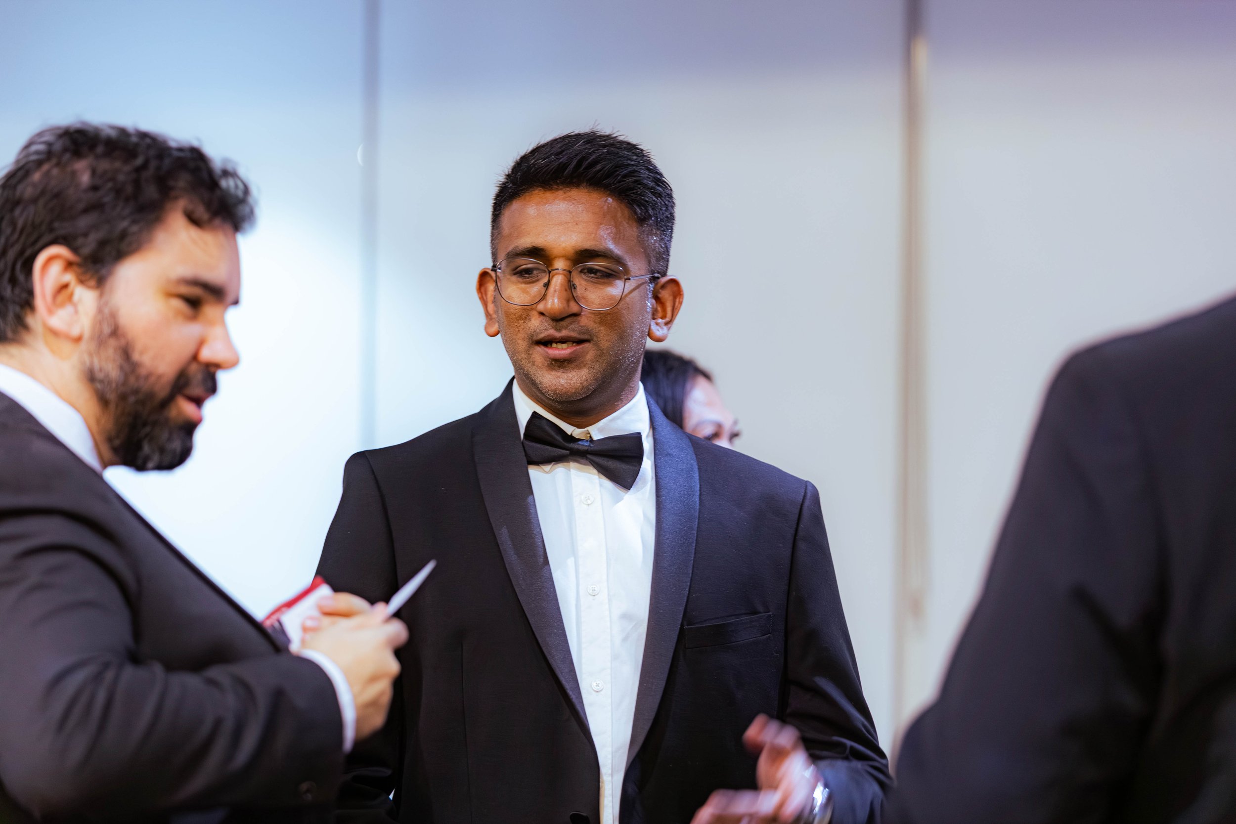 Three men in formal attire, two wearing tuxedos, are engaged in an indoor conversation. One man with glasses and a bowtie looks at the man on the right, who is mostly out of the frame.