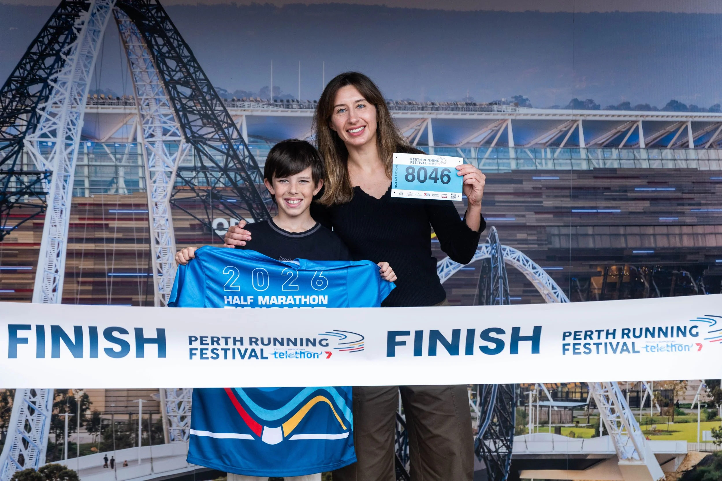 A woman and a young boy standing at the finish line of a half marathon event, holding a blue t-shirt. The woman is holding a race bib with the number 8046. They are smiling with a large Ferris wheel structure and a modern building in the background d