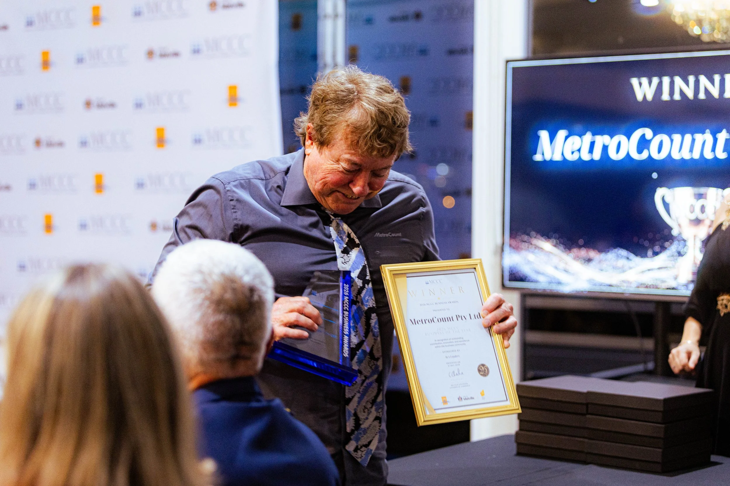 A man smiling while holding a framed certificate at an award ceremony. Several people are in the foreground, and a large screen displaying the MetroCount logo is visible behind him.
