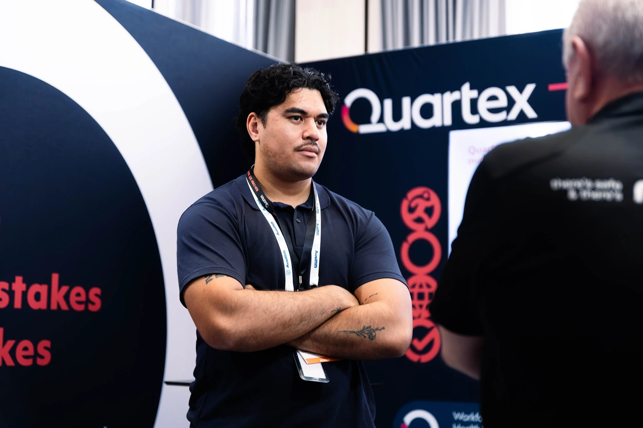 A young man with dark curly hair and tattoos on his arm is standing at a professional event, listening attentively to someone out of frame. He is wearing a navy blue polo shirt and a lanyard. Behind him is a black display with the word 'Qortex' and other text partially visible, indicating a booth at a conference or trade show.