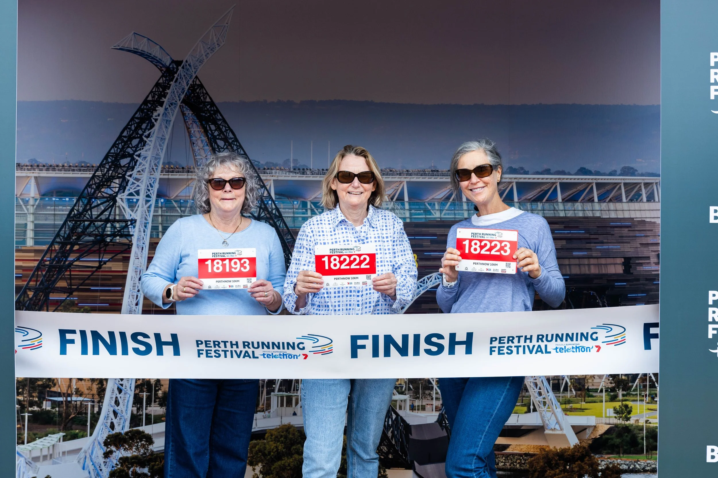 Three women wearing sunglasses and holding race bibs standing in front of a backdrop featuring the Perth cultural center with a large ferris wheel. The women are smiling, and the bibs read '18193', '18222', and '18223'. They are participating in the 